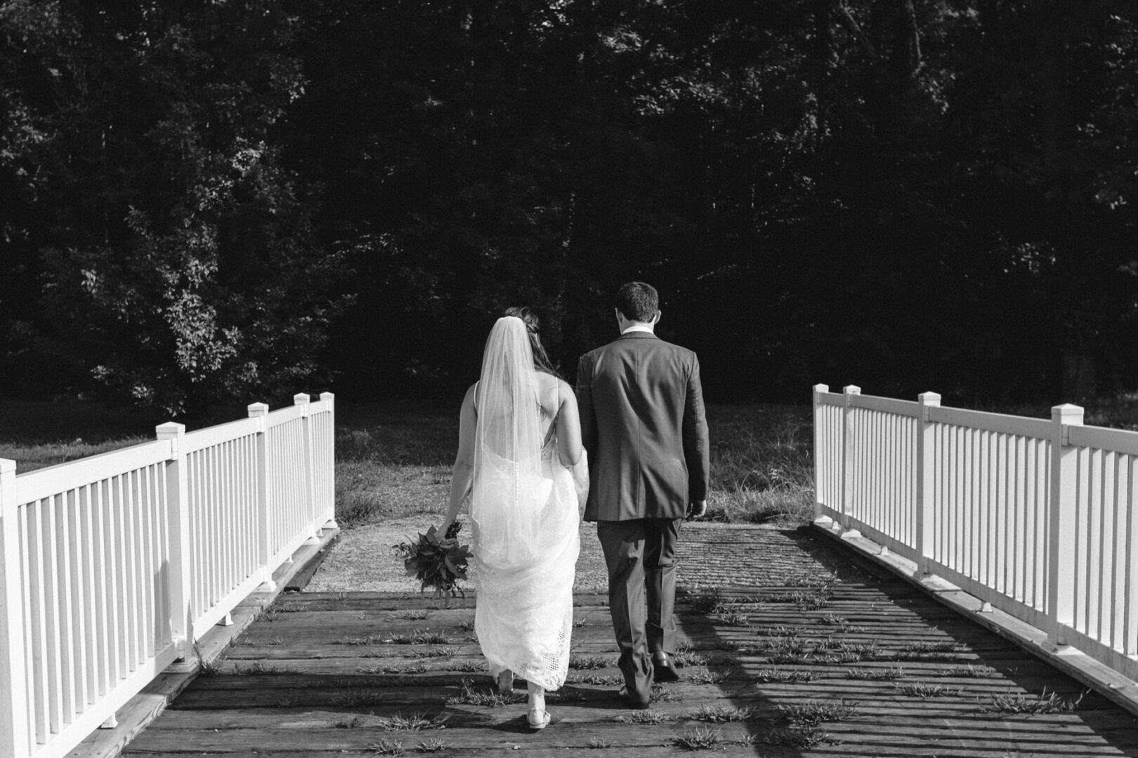 The bride and groom gracefully stroll across a wooden bridge adorned with white railings, embraced by a canopy of trees. This elegant black and white moment evokes the timeless charm found in Barn on Bridge wedding photos.