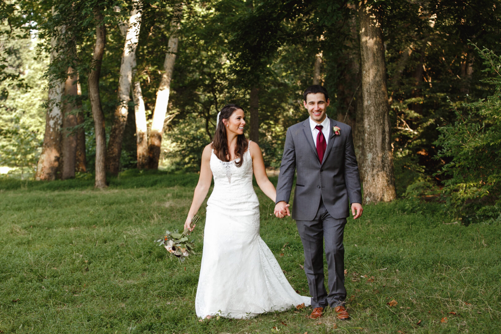 A bride and groom walk hand in hand on a grassy area surrounded by trees, capturing their love in charming Barn on Bridge wedding photos. The bride wears a white dress holding flowers, while the groom is stylishly dressed in a gray suit with a red tie.