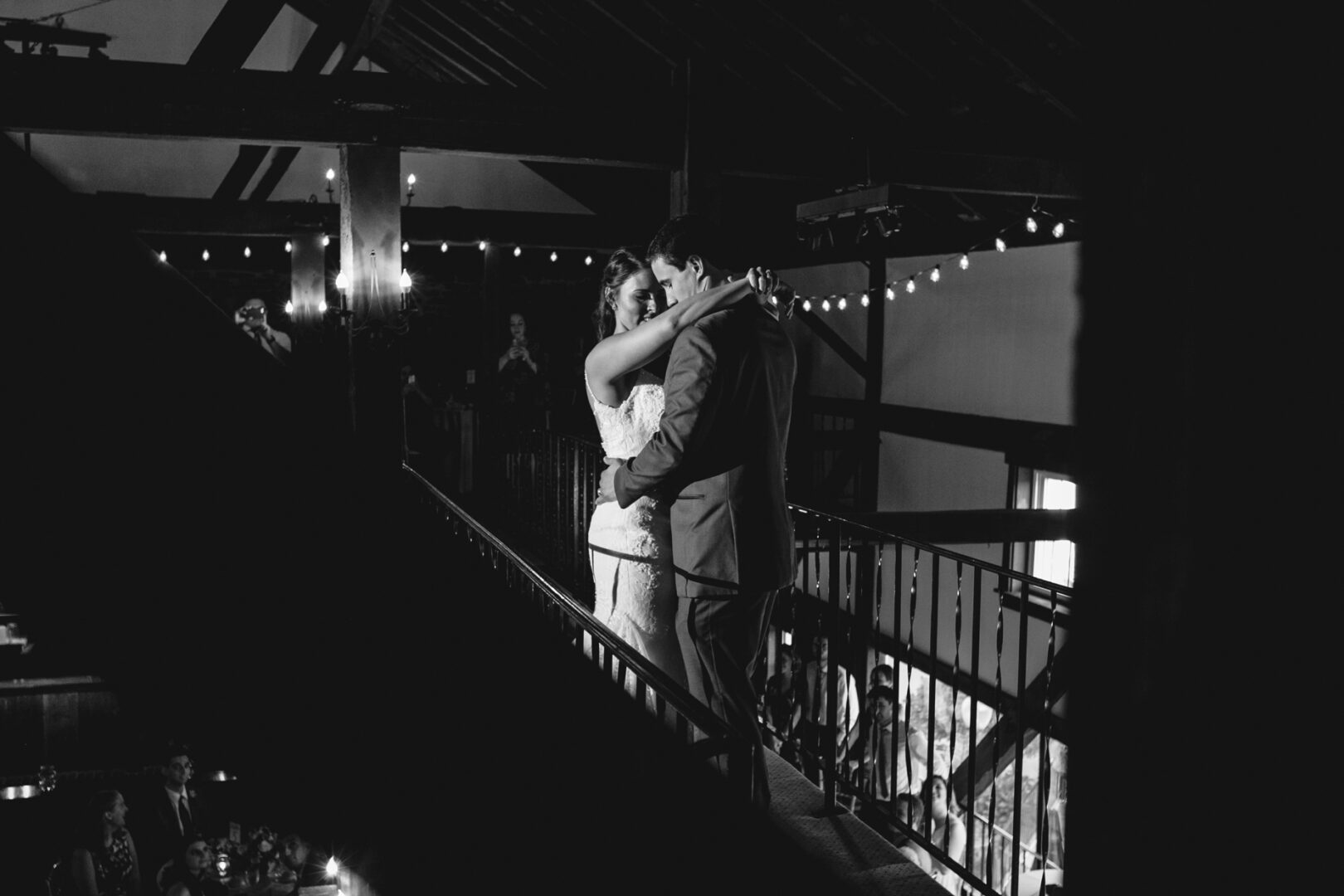 On a dimly lit balcony, a couple embraces during a dance, with string lights twinkling like stars. In the distance, memories are captured as timeless Barn on Bridge wedding photos.