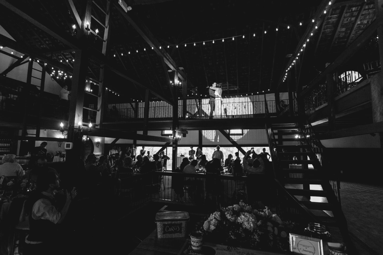 A black and white photo of a large indoor venue with a high ceiling, reminiscent of Barn on Bridge wedding photos. People are sitting and standing on a lower level, while two individuals gracefully dance on a raised platform.