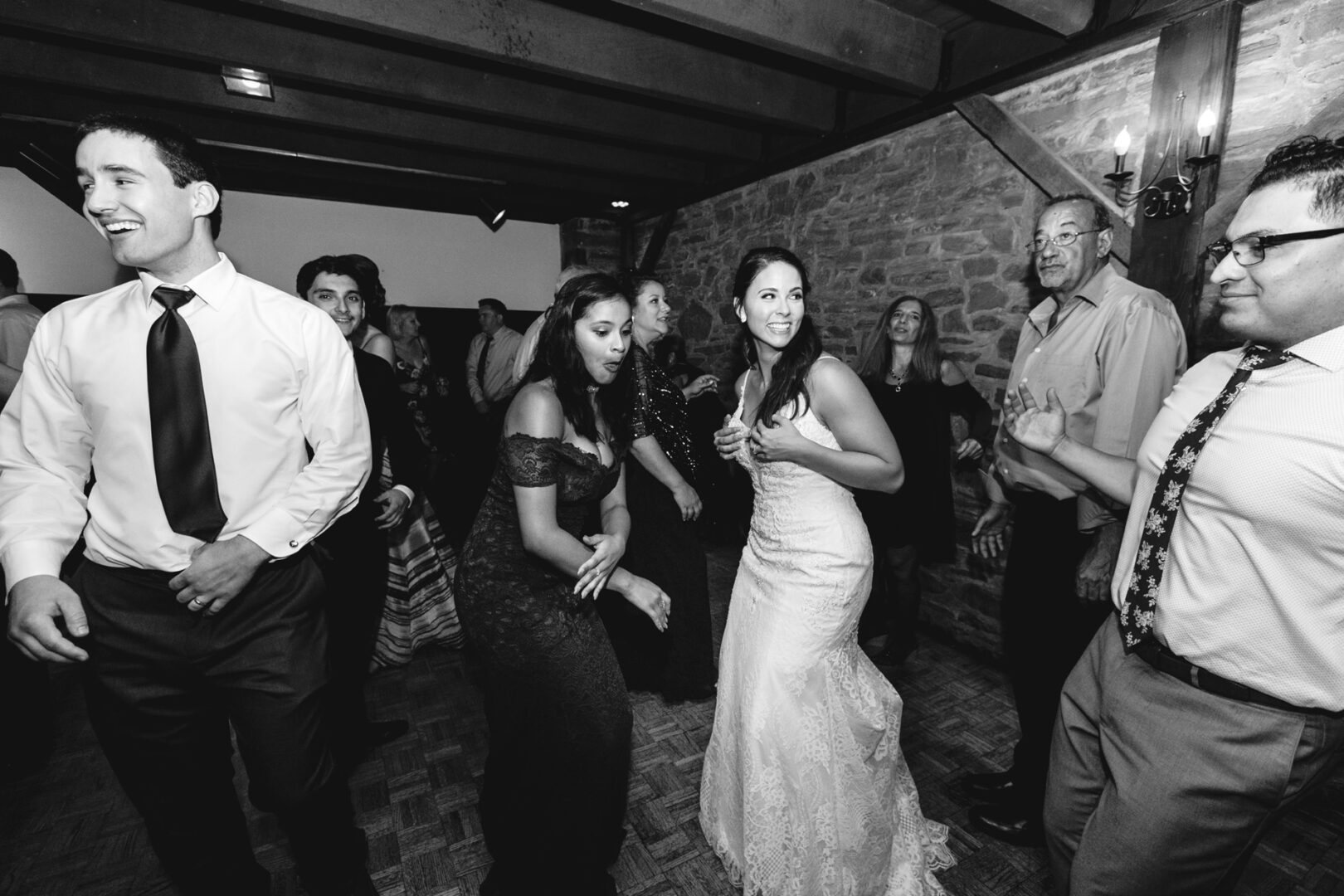 A group of people, including a bride in a white dress, joyfully dance at an indoor event. The charming setting at Barn on Bridge boasts rustic stone walls and wooden beams, adding timeless elegance to the wedding photos.