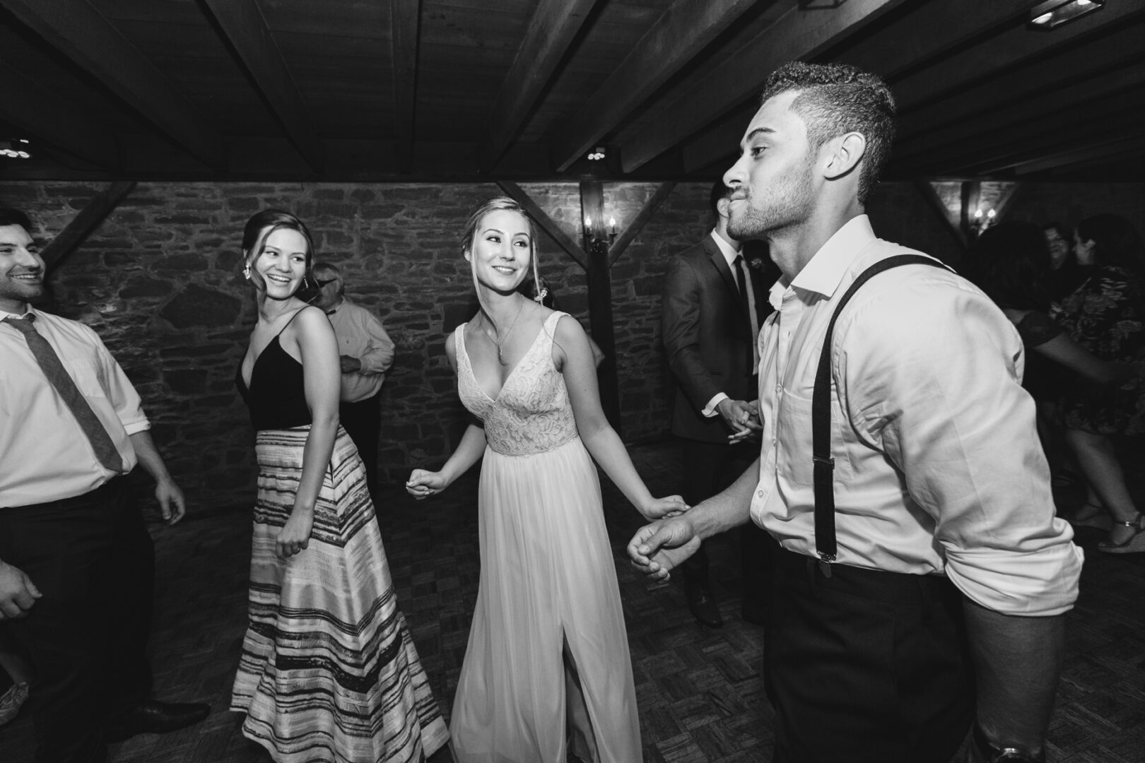 In a dimly lit room with stone walls, perhaps from the popular Barn on Bridge Wedding Photos collection, people dance joyfully. A woman in a light dress smiles, holding hands with a man in shirt and suspenders as others twirl nearby.