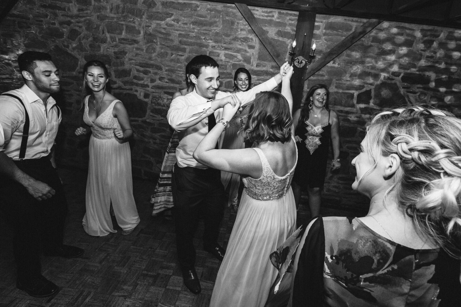 A group of people dancing energetically in a room with a stone wall, reminiscent of the charming Barn on Bridge wedding photos. The focus is on a couple holding hands, surrounded by others in formal attire, all exuding joy and elegance.