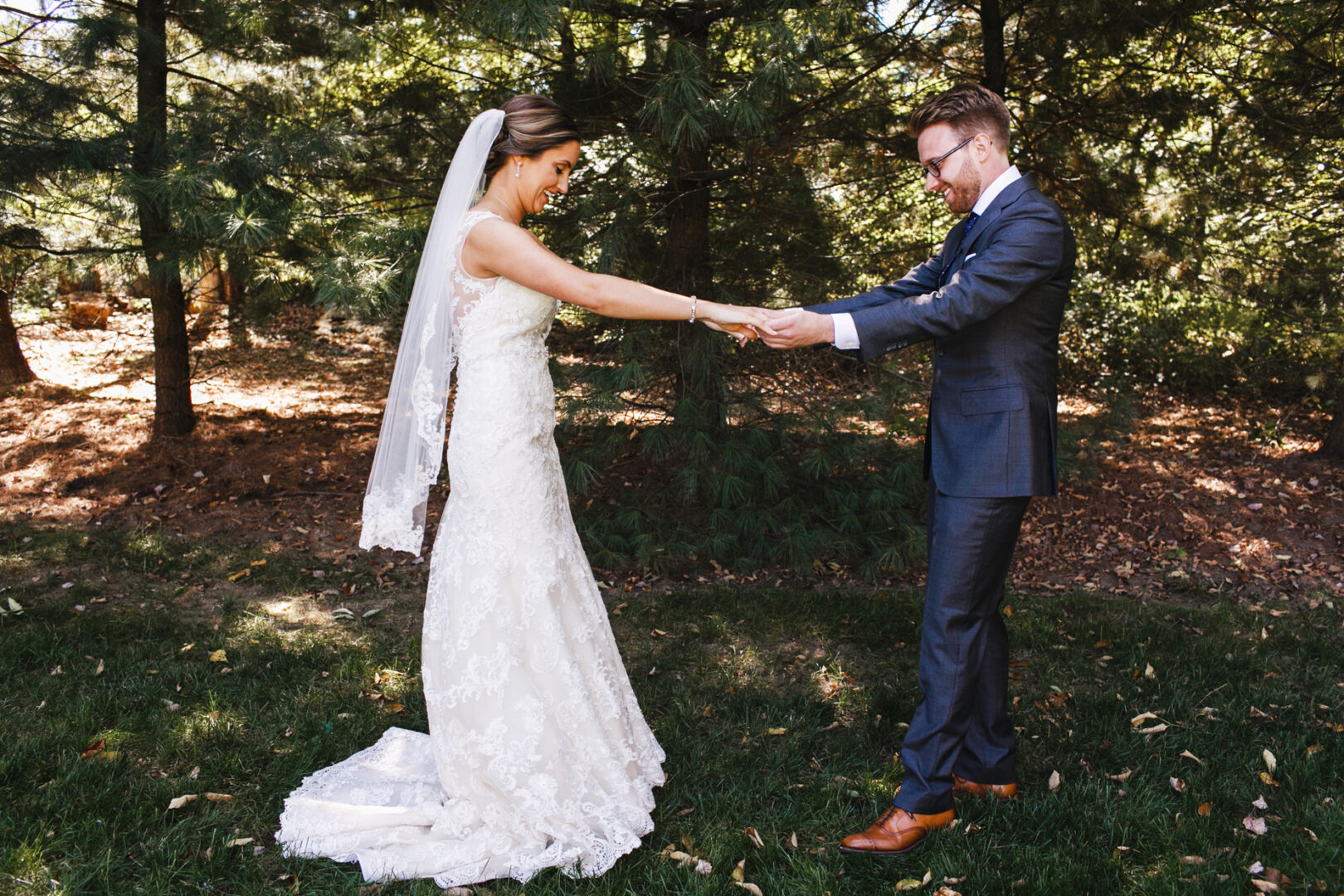 The bride and groom, beaming with joy, hold hands amid the lush trees at The Manor House at Commonwealth Wedding. She dazzles in a white gown with a veil, while he stands elegantly in a dark suit and brown shoes.
