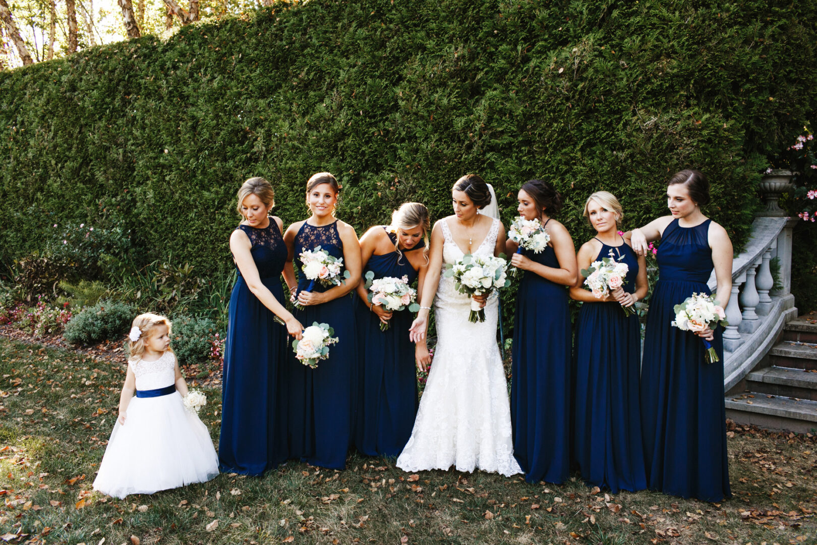 Bride and six bridesmaids in navy blue dresses pose for a photo next to a young flower girl in a white dress. They are outdoors at The Manor House at Commonwealth Wedding, standing before a lush green hedge on a sunny day.