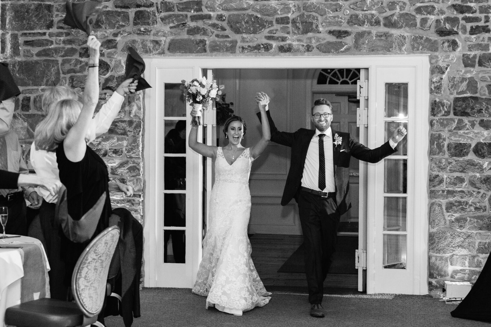Newlywed couple enters a room at The Manor House at Commonwealth Wedding, smiling and hand-in-hand, as guests cheer and wave napkins. Stone wall and glass doors provide a charming backdrop.