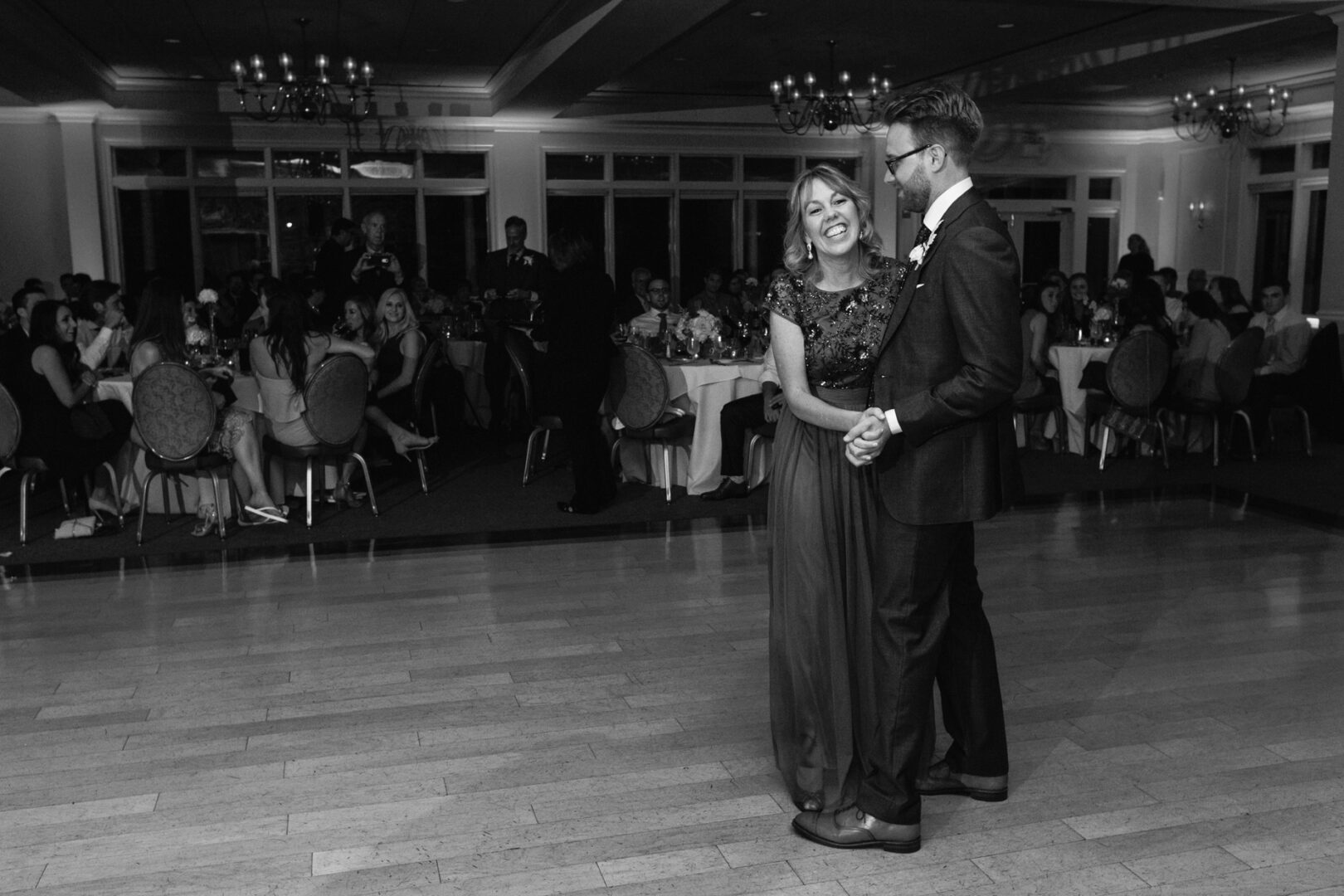 At The Manor House at Commonwealth Wedding, a couple elegantly dances together on a wooden floor, while guests seated at tables in the background admire the enchanting scene.