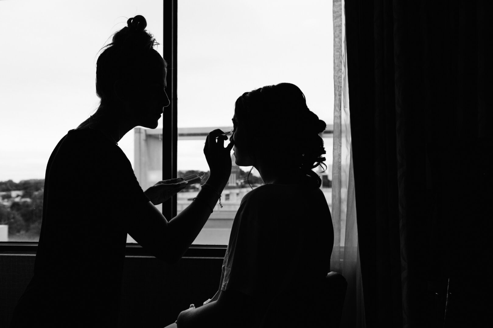 Silhouette of a makeup artist meticulously applying makeup to a seated person, with the elegance reminiscent of preparations for a Rockwood Park Carriage House Wedding, all framed by the soft light streaming through the window.