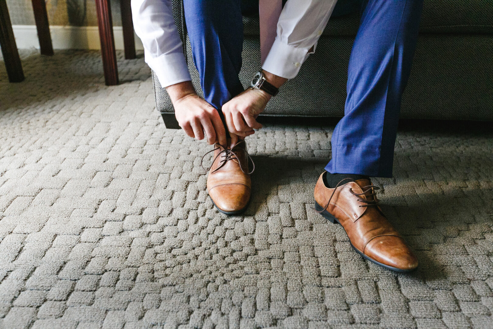 Amid the elegance reminiscent of a Rockwood Park Carriage House Wedding, a person in blue pants and a white shirt ties brown dress shoes while seated on a sofa.