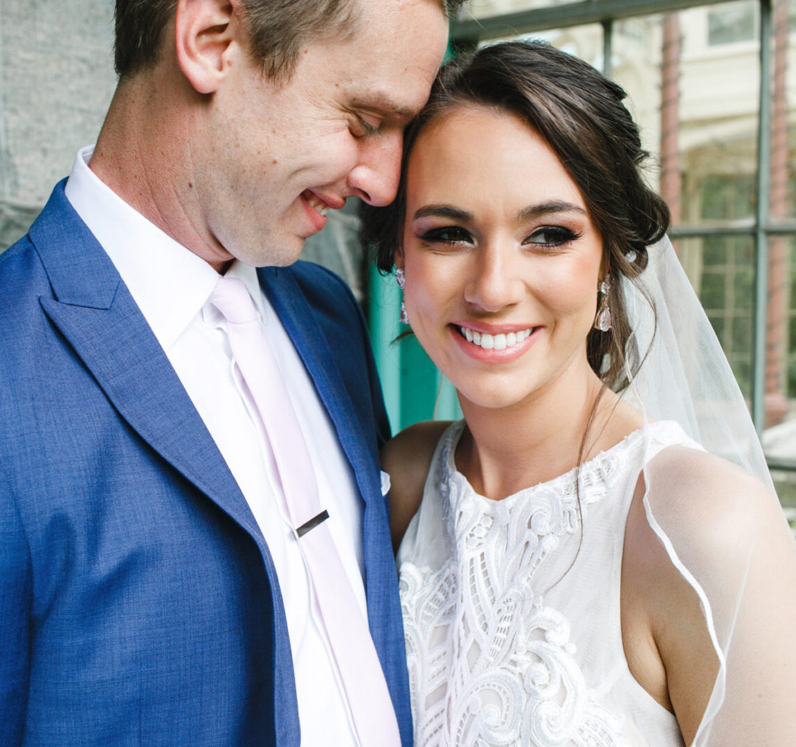 A smiling couple stands close together at the Rockwood Park Carriage House. The man is wearing a blue suit, and the woman is in a white dress with lace details, near a stone wall and window, capturing the essence of their perfect wedding day.
