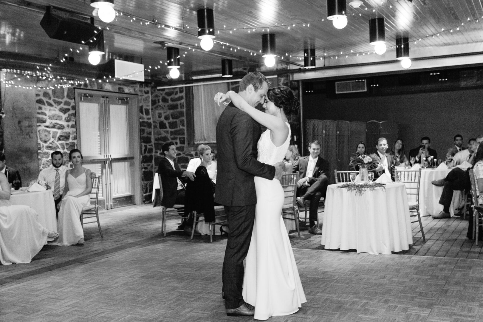 A bride and groom share a dance in the warmly lit Rockwood Park Carriage House, surrounded by seated guests and elegantly decorated tables.