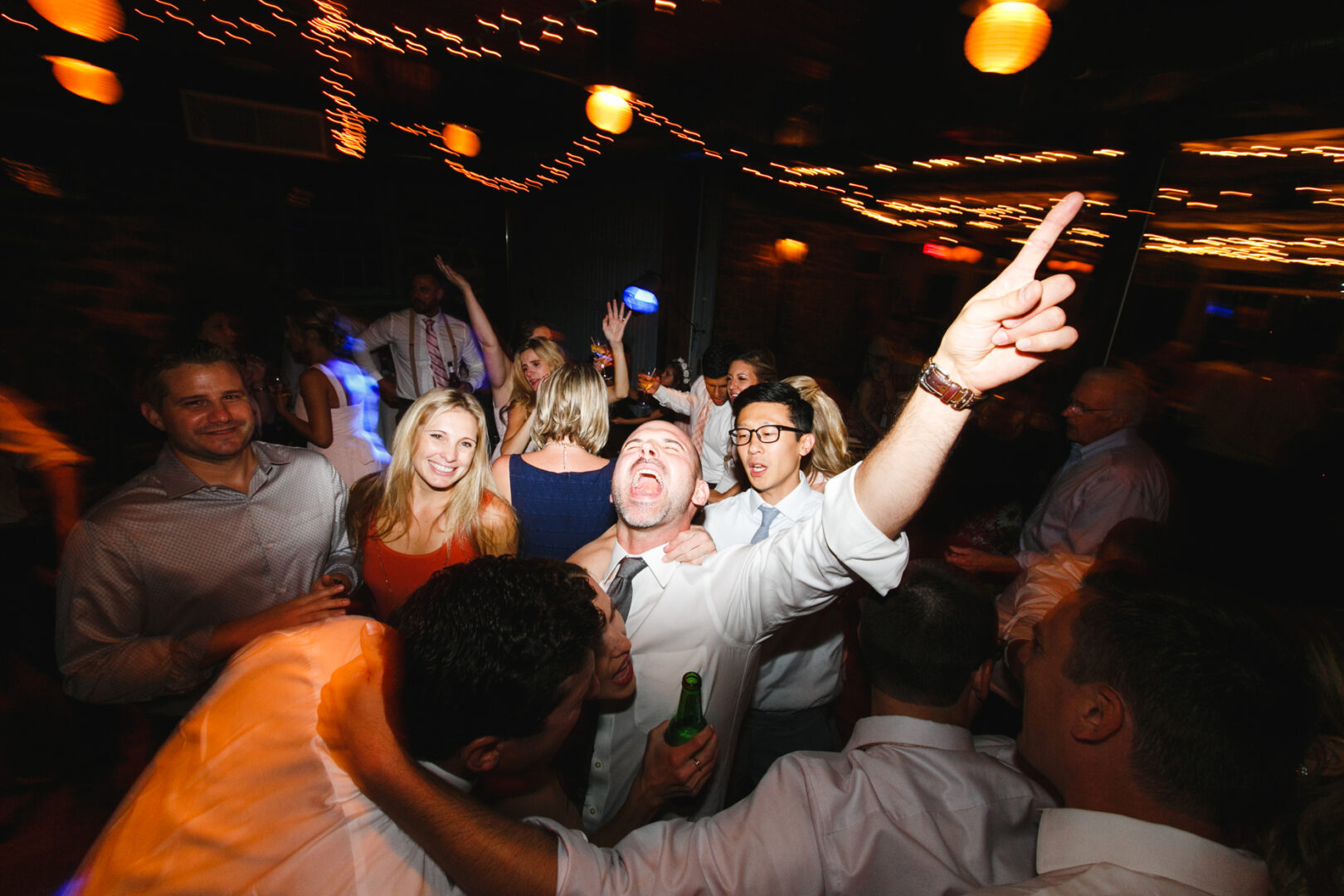 A lively indoor celebration at the Rockwood Park Carriage House Wedding features a group dancing amid vibrant lights and decorations. One person in the foreground gestures energetically, capturing the joyous spirit of the event.