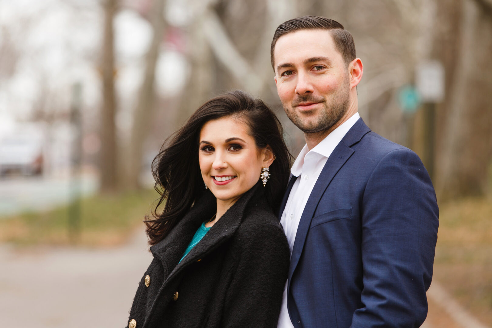 A man in a blue suit and a woman in a black coat stand side by side outdoors, capturing the essence of their NYC engagement session with trees artistically blurred in the background.