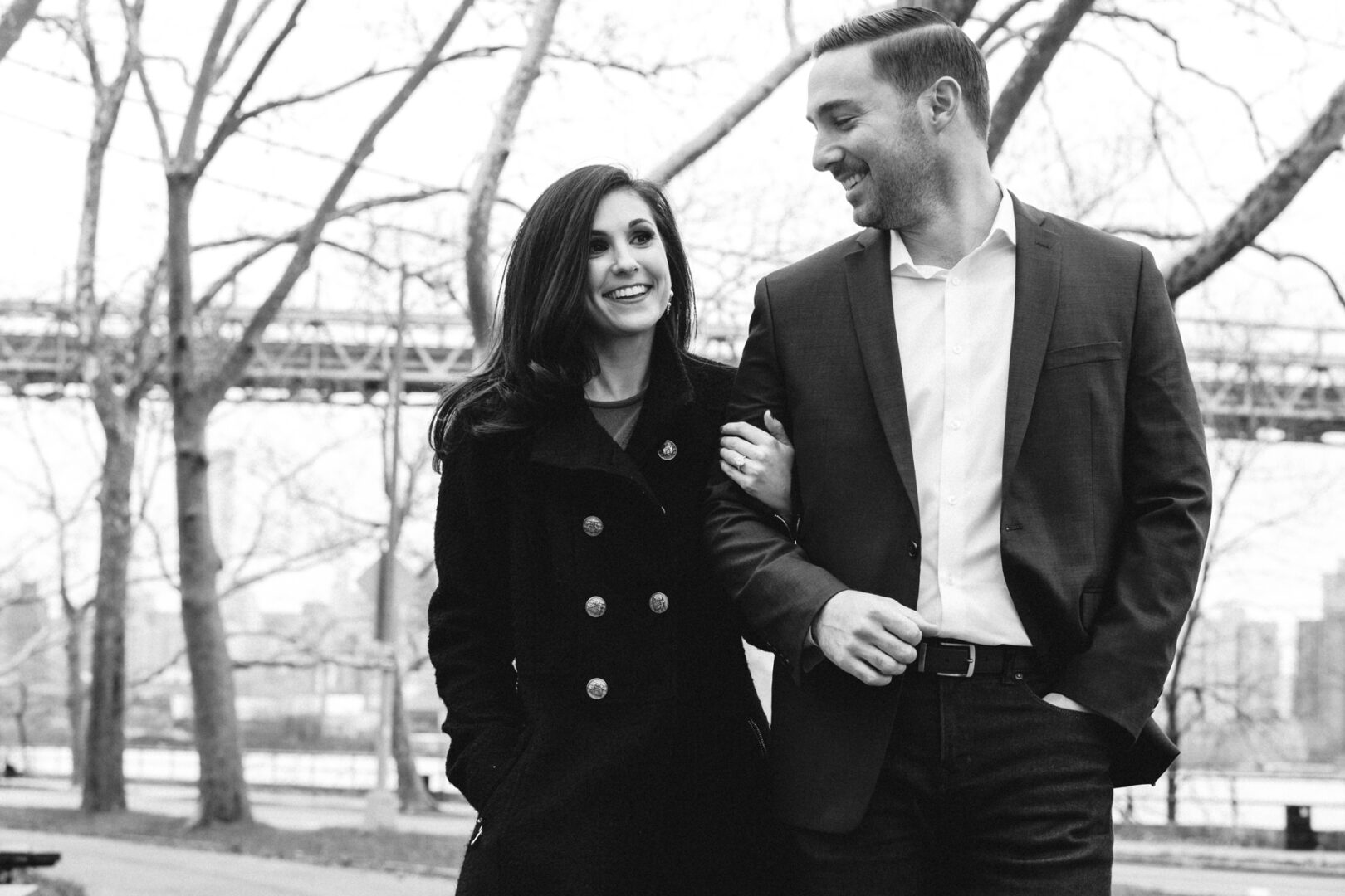 During their NYC engagement session, a man in a suit and a woman in a coat stroll arm in arm through the winter park, framed by bare trees and an elegant bridge.