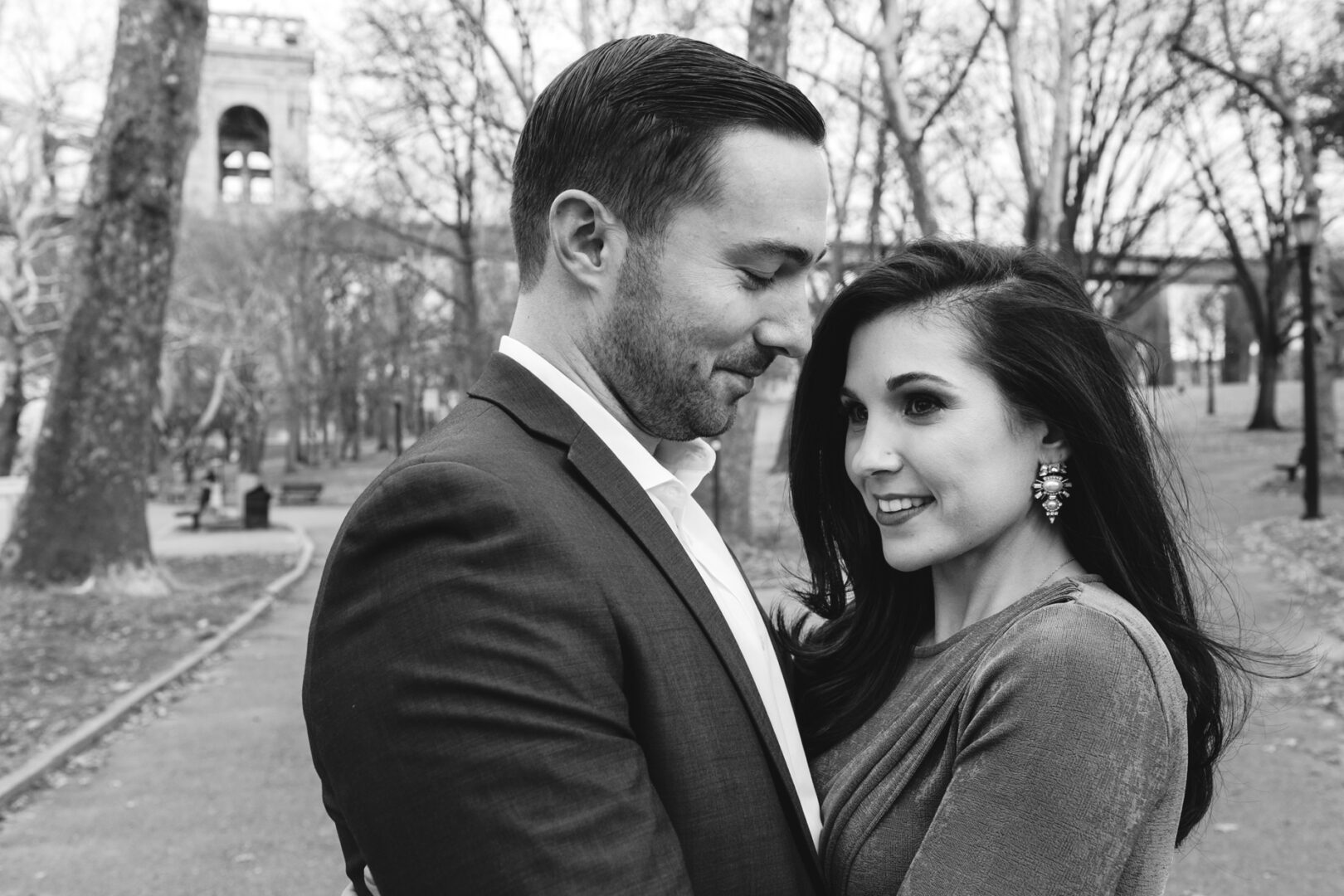 A couple stands closely together in a park, surrounded by leafless trees and paths. The man wears a suit, and the woman dons a dress with large earrings. This NYC engagement session is captured in an elegant black and white photo.
