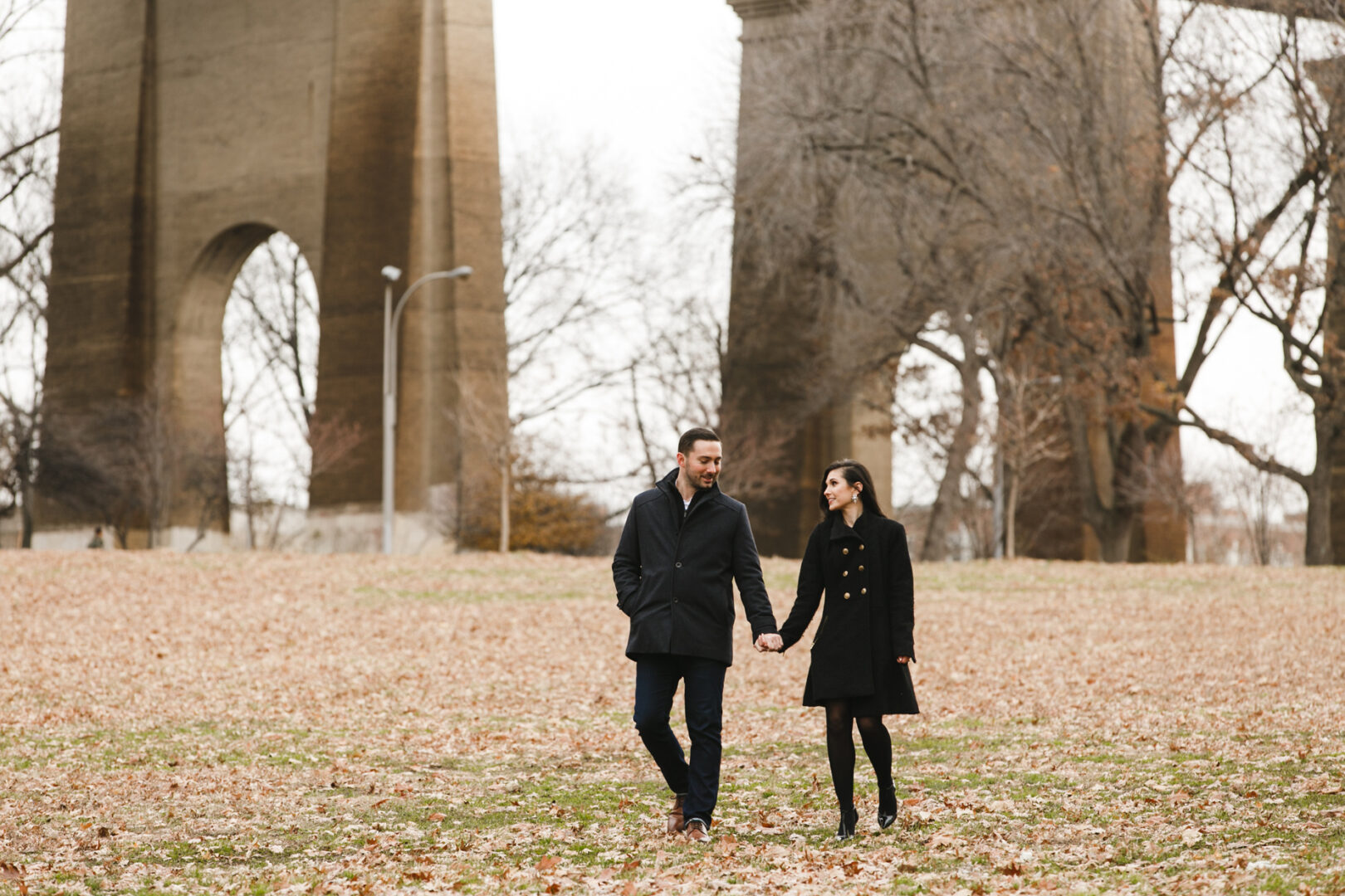 A couple holding hands wanders through a park in an NYC engagement session, with tall stone structures framed against an overcast sky.