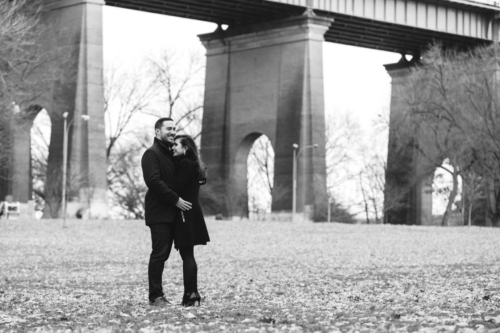 A couple stands closely together on the leaf-covered ground in front of a large bridge with towering pillars, capturing the essence of an NYC engagement session. Trees without leaves frame the backdrop in this evocative black-and-white image.