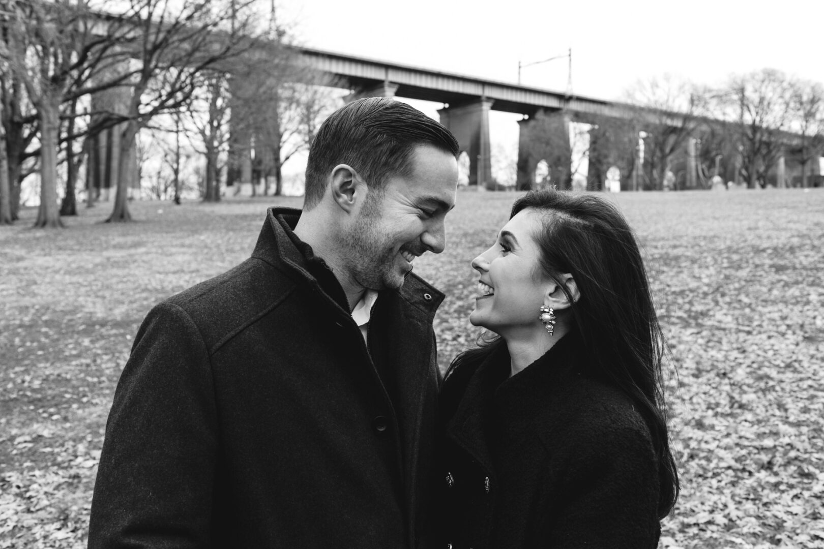 A man and woman smiling at each other in a park during their NYC engagement session, with a bridge in the background, surrounded by leafless trees.