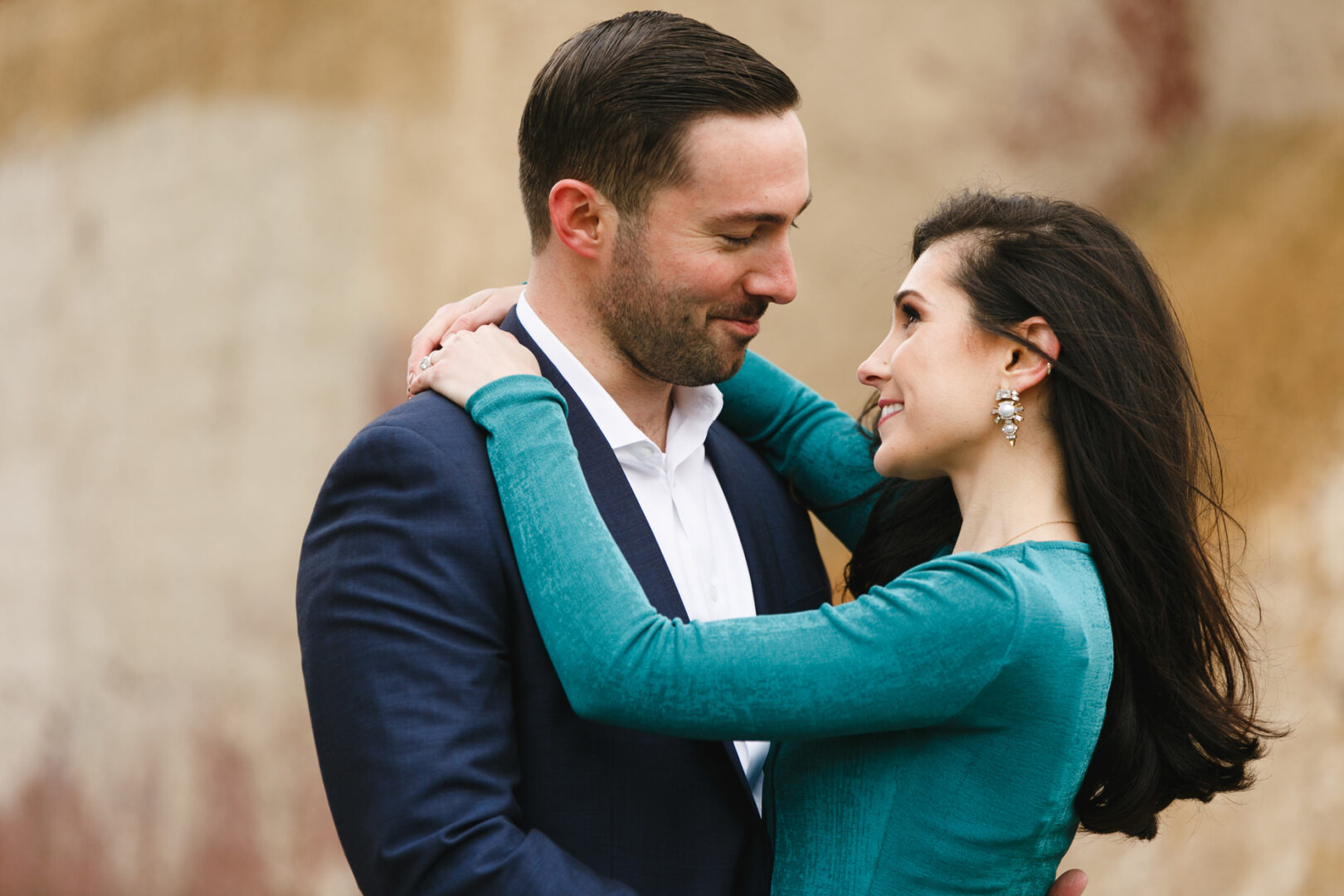 A couple embraces closely, both smiling gently, capturing a timeless moment during their NYC engagement session. The man is in a blue suit, while the woman dazzles in a teal dress with earrings against a neutral background.