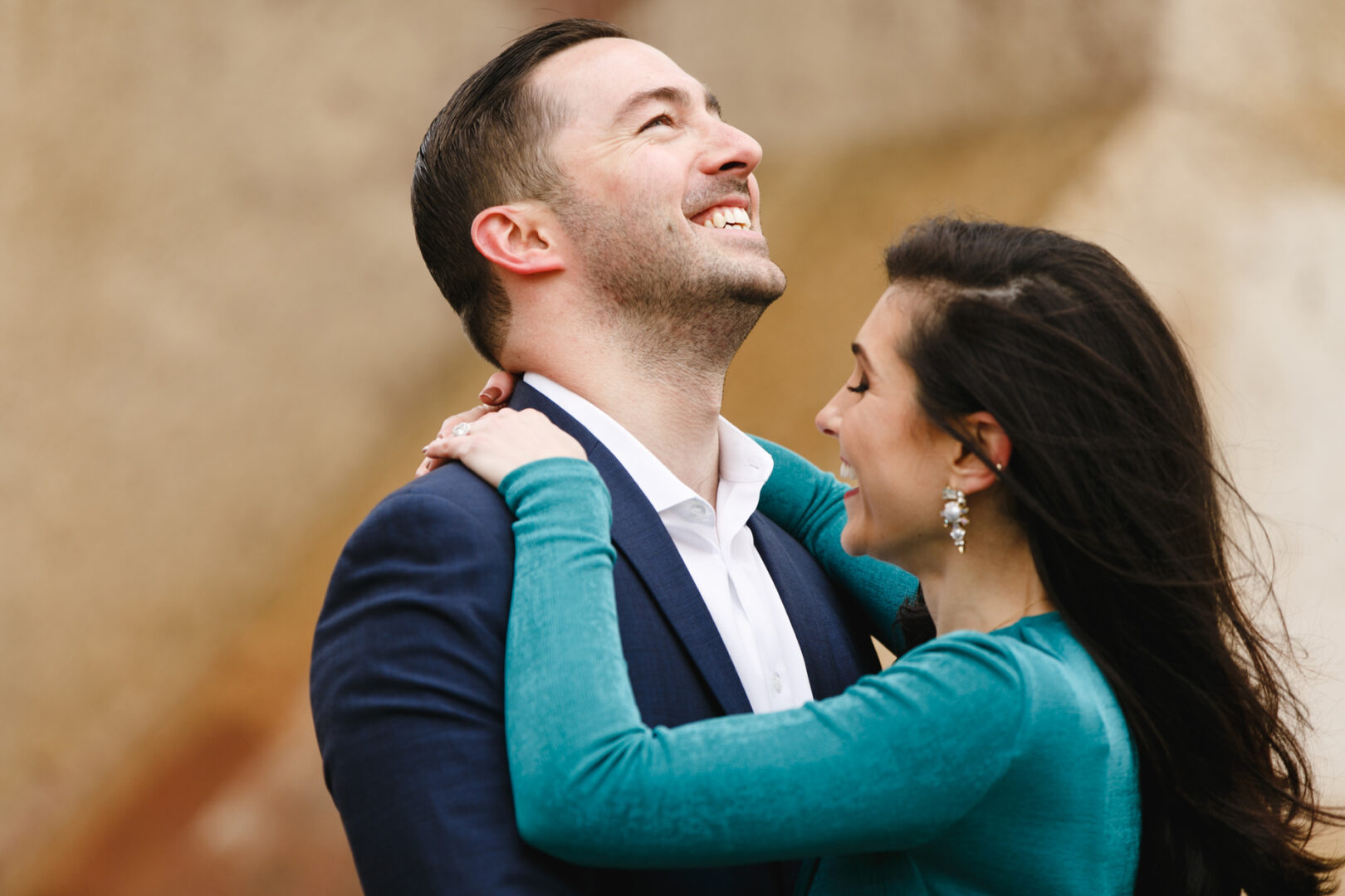 A man and woman are smiling and embracing, with the woman's hands on the man's shoulders, capturing their joy during an NYC engagement session. The background is blurred and neutral-toned.