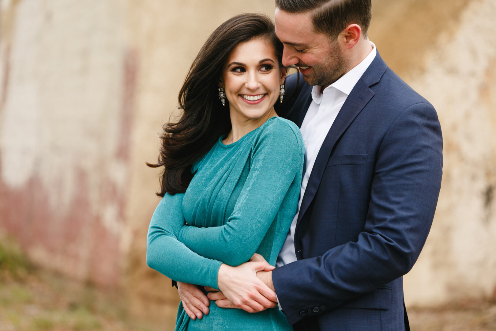 Against a textured, muted wall, the couple embraces during their NYC engagement session. The woman in a teal dress smiles warmly as the man, in a navy suit, gazes at her affectionately.