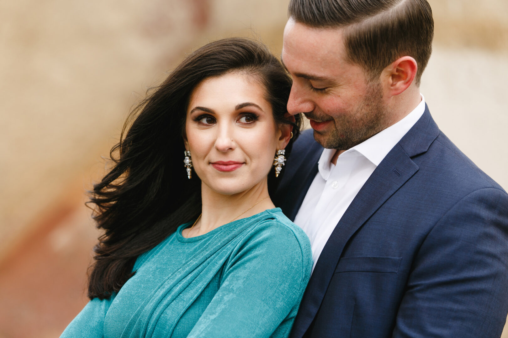 In this NYC engagement session, a man in a blue suit embraces a woman in a teal dress. Both have dark hair. The woman looks to her right, while the man gazes at her with a slight smile amidst the blurred city background.