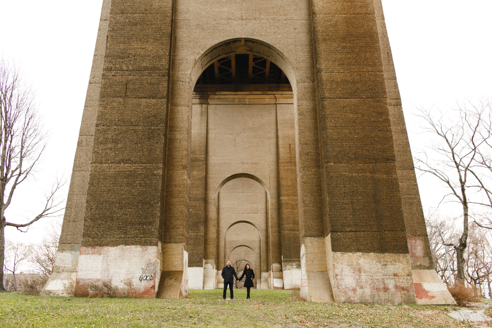 Two people stroll beneath a sweeping concrete bridge with multiple arches, the perfect NYC engagement session backdrop. Lush grass and trees add a touch of nature's charm to the urban scene.