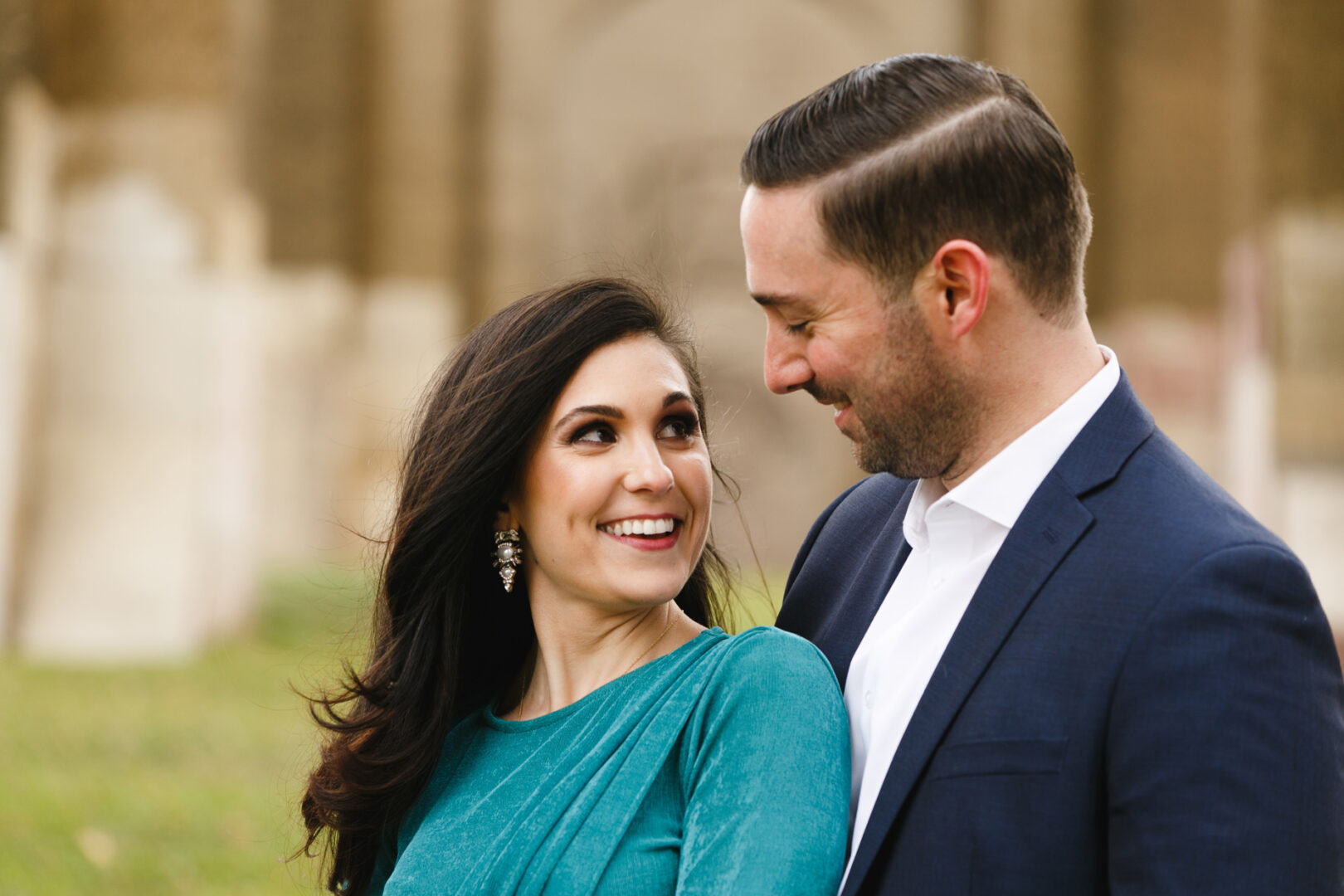A man in a blue suit and a woman in a green dress smile at each other outdoors, capturing the joy of their NYC engagement session.