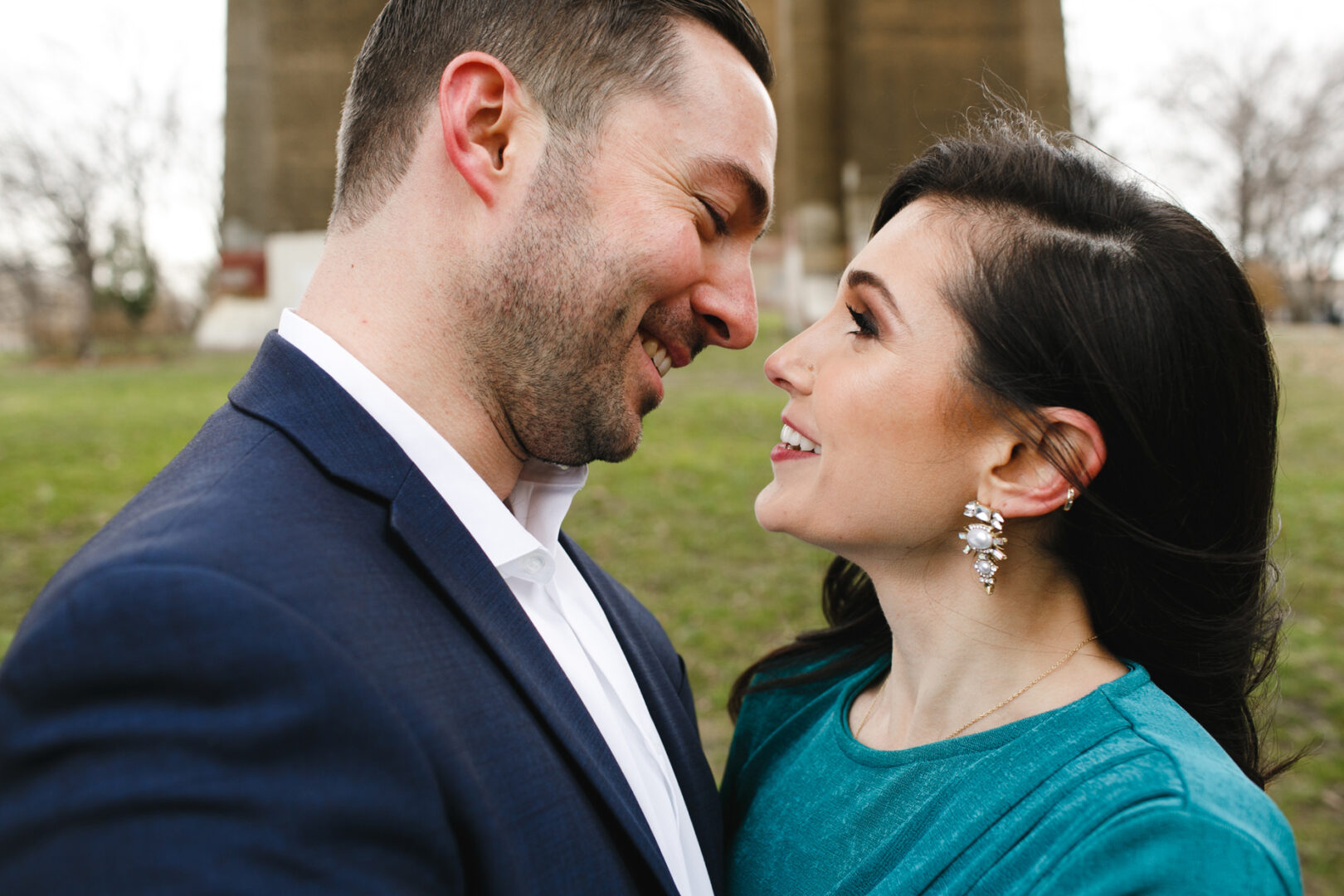 A man and woman are smiling at each other outdoors during their NYC engagement session. She wears a teal top and statement earrings, while he is in a dark suit. Standing closely with blurred trees in the background, their joy is as vibrant as the city they love.