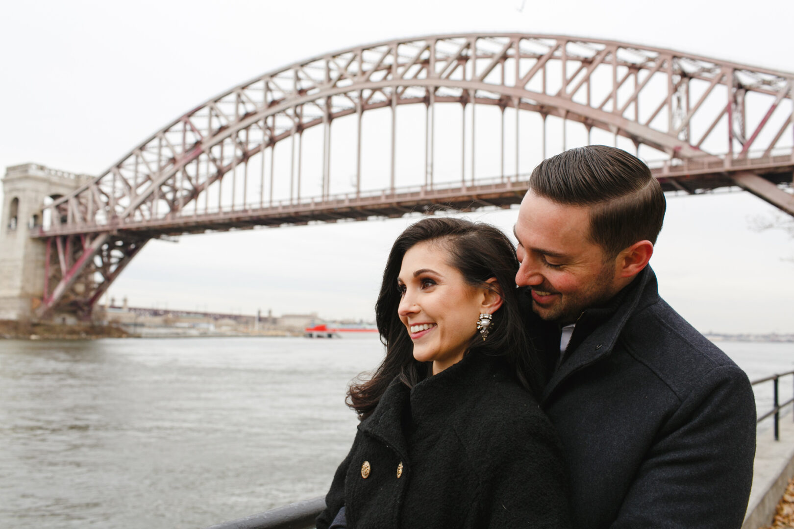 The couple beams with joy by the river, the iconic arch bridge framing their NYC engagement session perfectly.