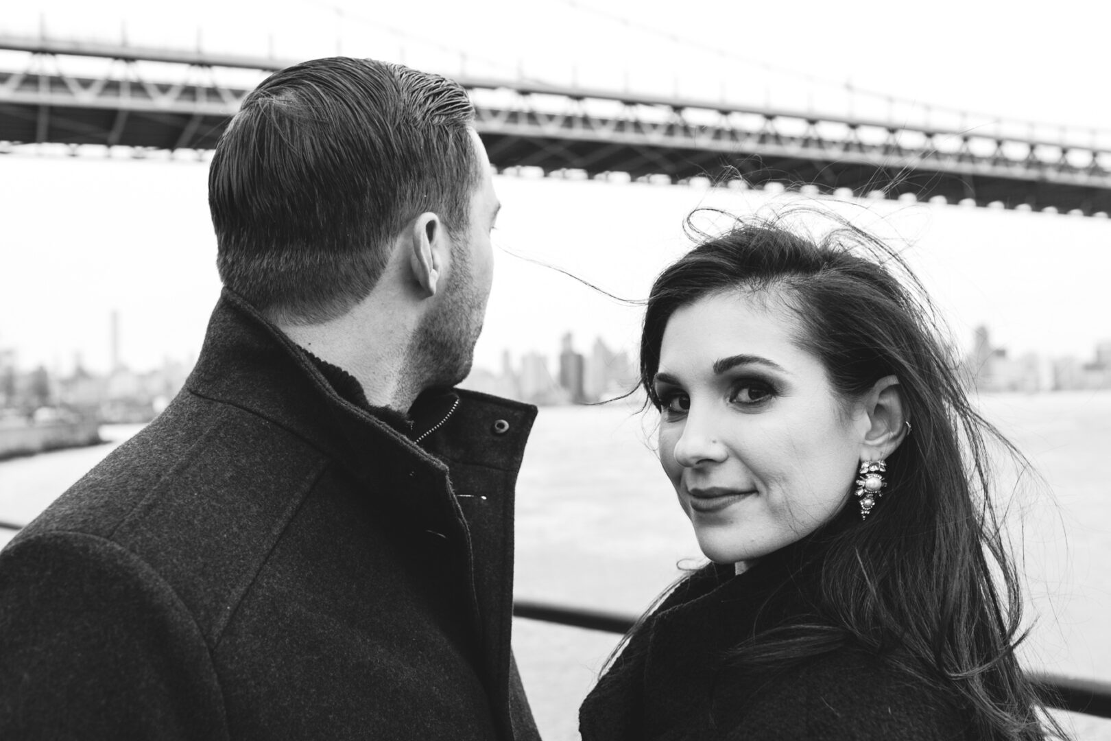 A woman gazes at the camera, a man beside her, both donning coats, with a sweeping bridge over the river behind them. Captured in timeless black and white, this NYC engagement session embodies classic romance.