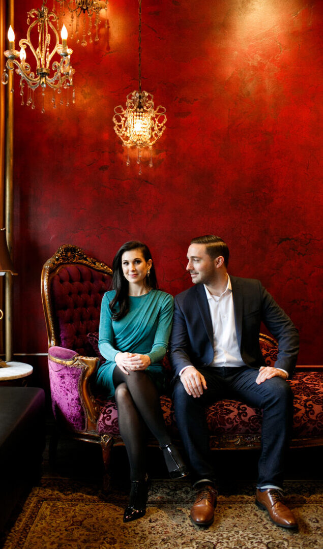 A man and woman sit on a vintage red and purple couch against a red wall, ornate lights casting a warm glow, as they face each other. This intimate setting sets the perfect backdrop for their NYC engagement session.