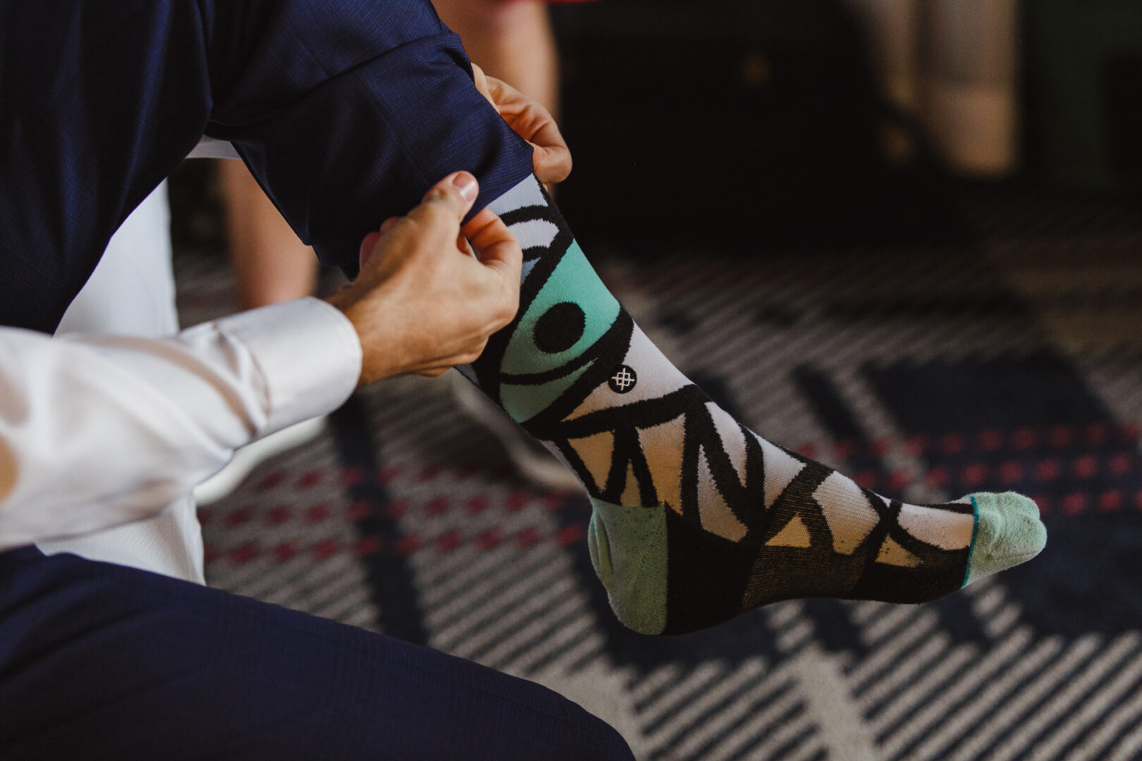 A person in a white shirt and dark pants adjusts a colorful sock with a geometric pattern while sitting on a patterned carpet at the Horticultural Center wedding.
