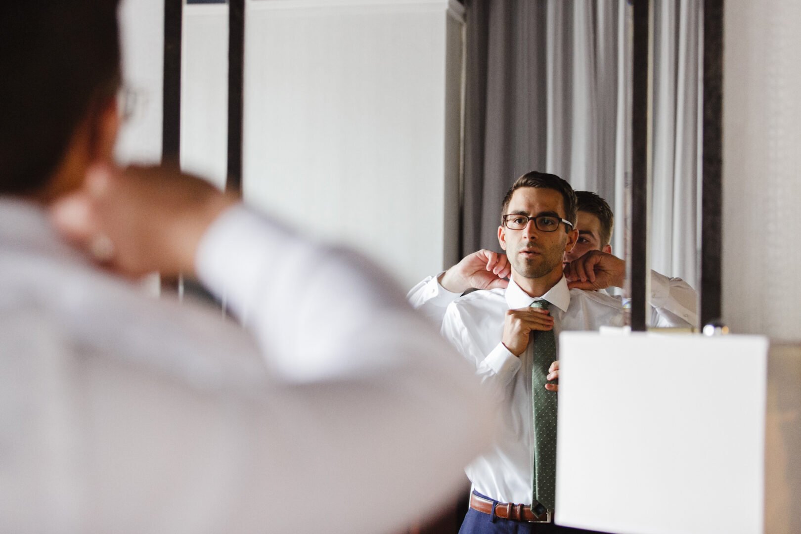 A man adjusts his tie in front of a mirror, wearing a white shirt and a green tie, his focused expression reflecting the anticipation of a Horticultural Center wedding.