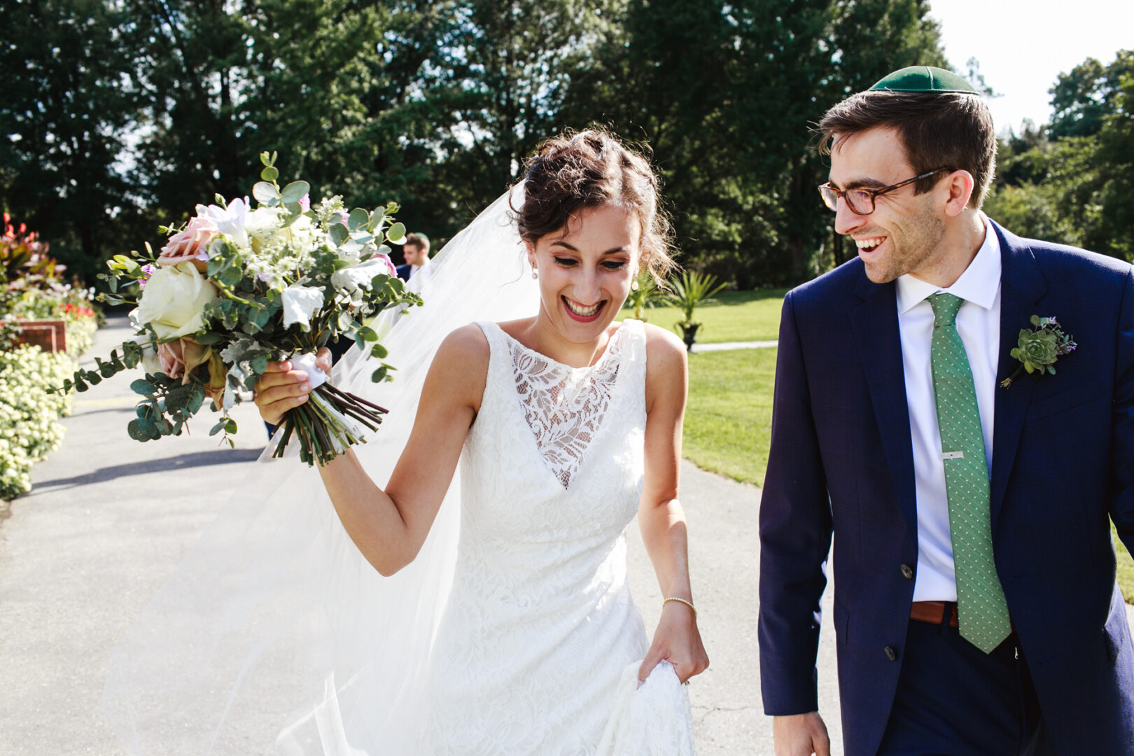 A bride and groom walk joyfully outdoors at the Horticultural Center. The bride holds a bouquet, wearing a white dress and veil, while the groom dons a dark suit with a green tie. They are surrounded by lush greenery and sunlight.