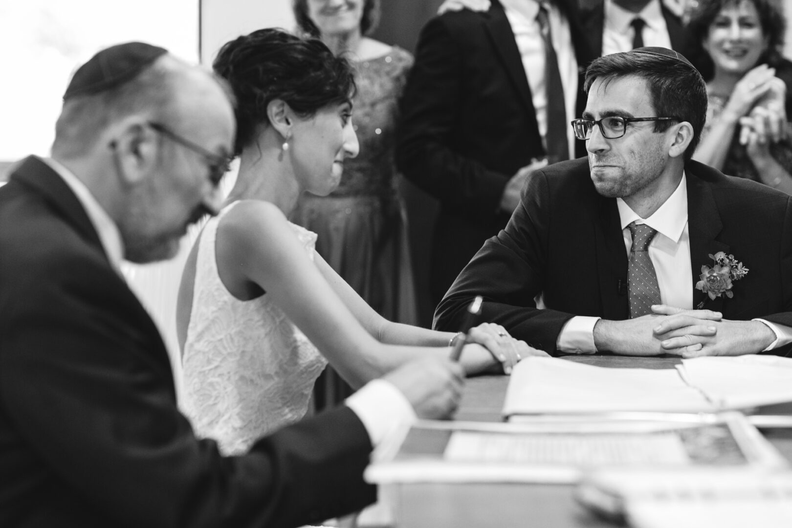 A bride and groom sit across from an officiant signing documents, surrounded by guests at their charming Horticultural Center wedding, captured in a timeless black and white photo.