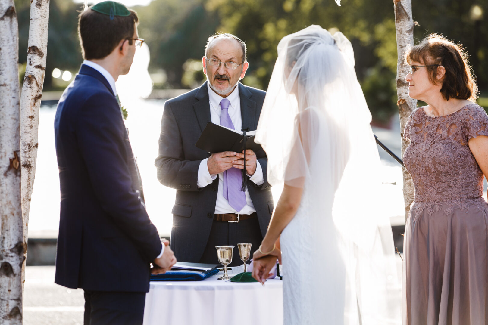 A wedding ceremony unfolds at the Horticultural Center, with a couple standing before an officiant. The groom wears a suit; the bride is in a white dress with a veil. Two goblets are elegantly placed on a table nearby amidst lush greenery.