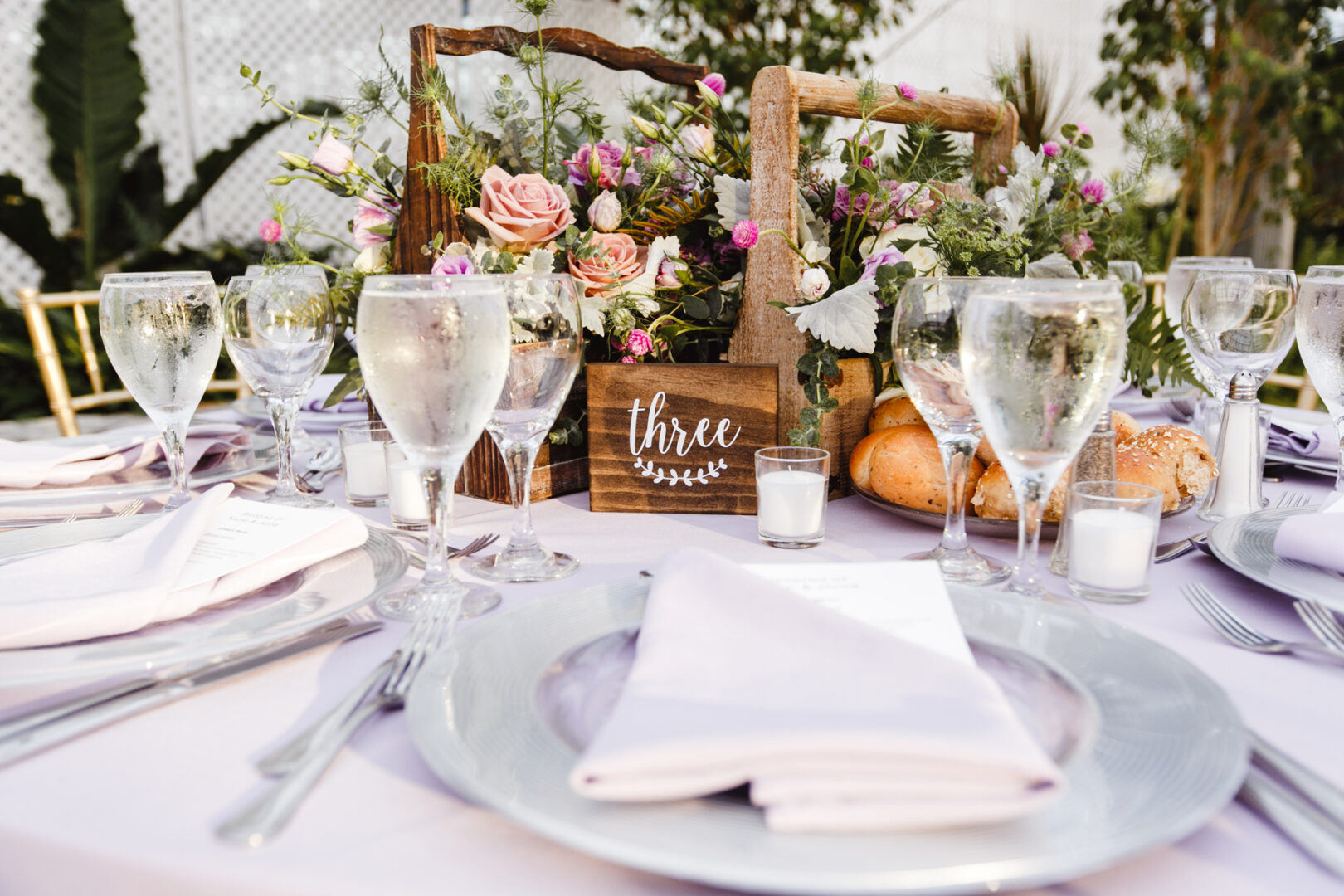A decorated table setup with plates, glasses, and napkins exudes rustic charm at this Horticultural Center wedding. A wooden centerpiece with flowers and a "three" sign is surrounded by candles and bread rolls, creating an inviting ambiance for guests.