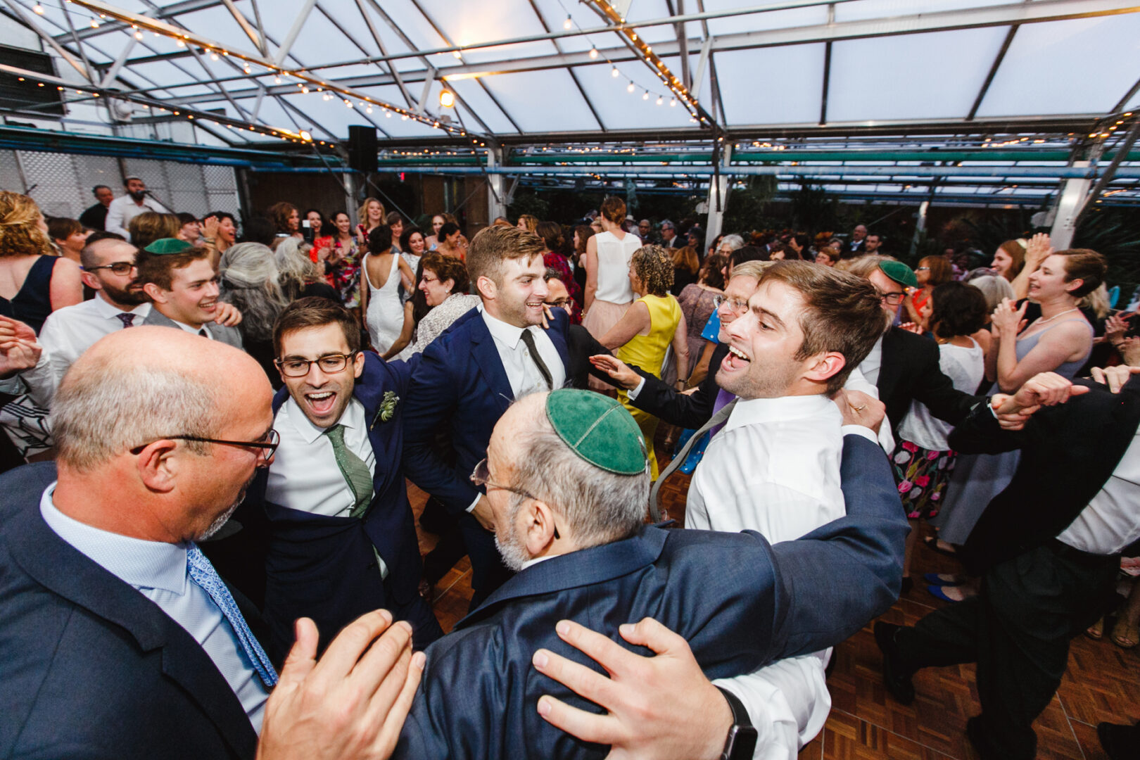 At a lively Horticultural Center wedding, a group of men in suits and ties laugh and dance joyously in a circle, surrounded by a bustling crowd of celebrants.