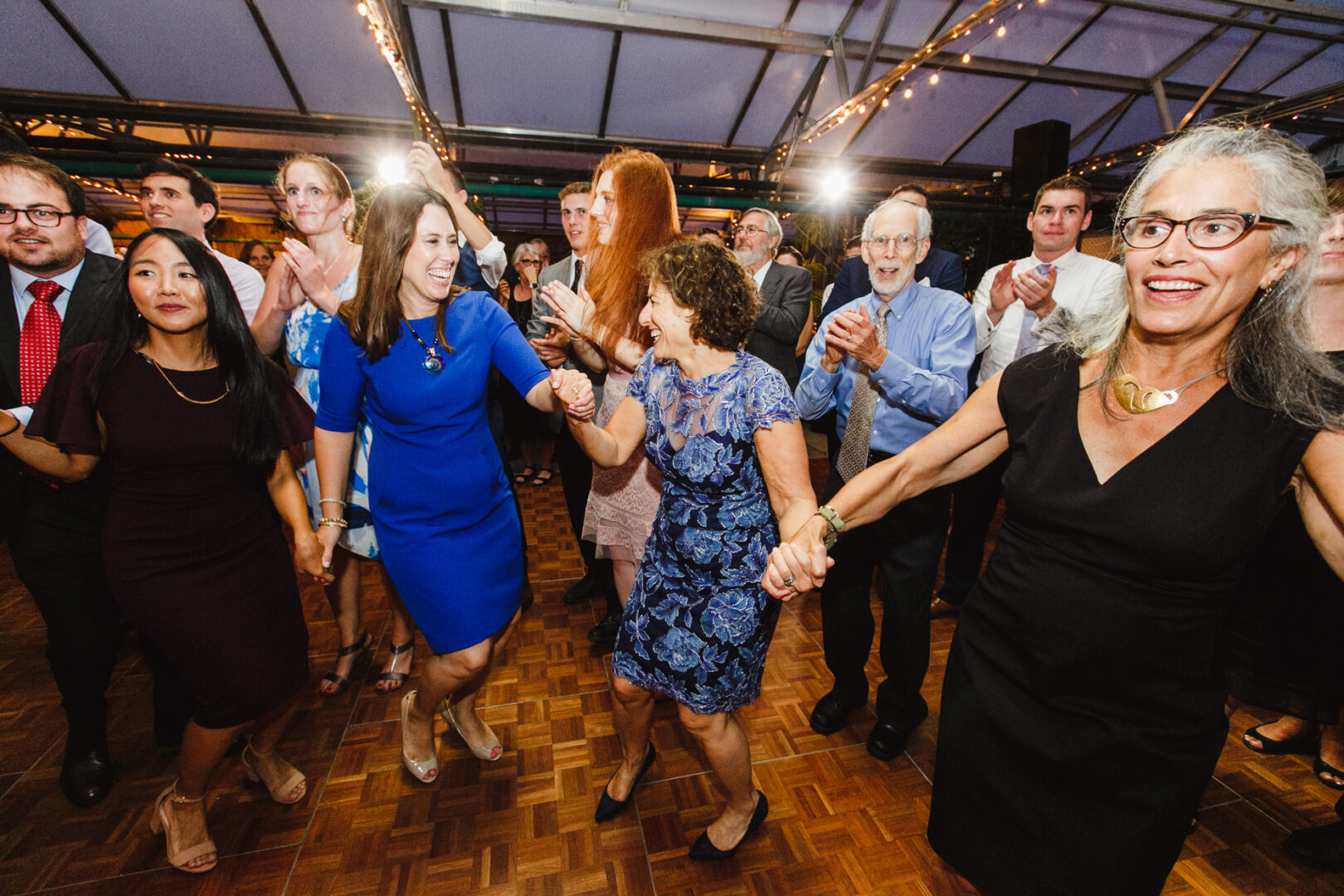 A group of people in formal attire are dancing and holding hands under event lighting on a wooden floor, celebrating a joyous occasion perhaps at a Horticultural Center wedding.