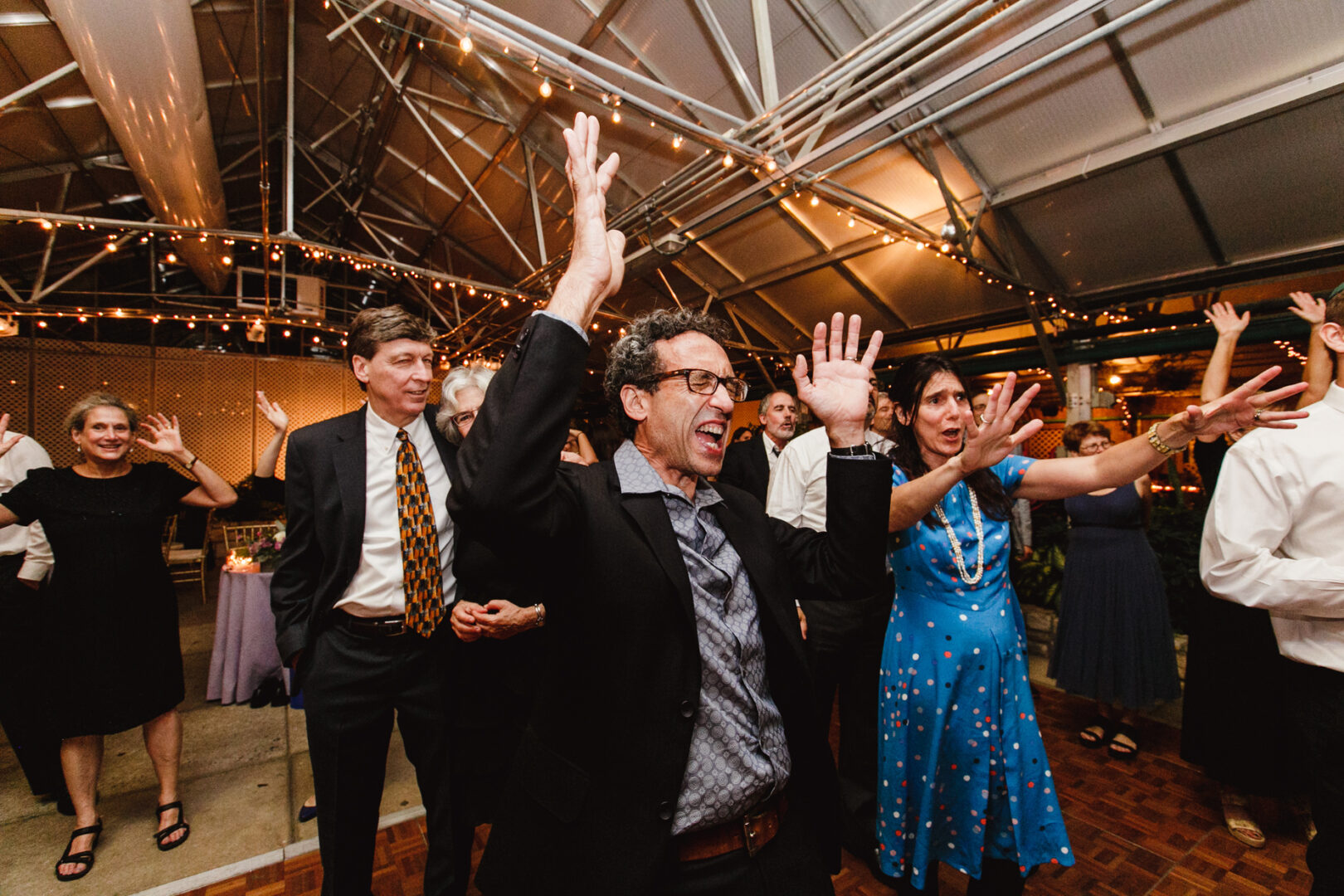 People in formal attire dance enthusiastically with raised hands at a warmly lit Horticultural Center wedding, under a high ceiling.