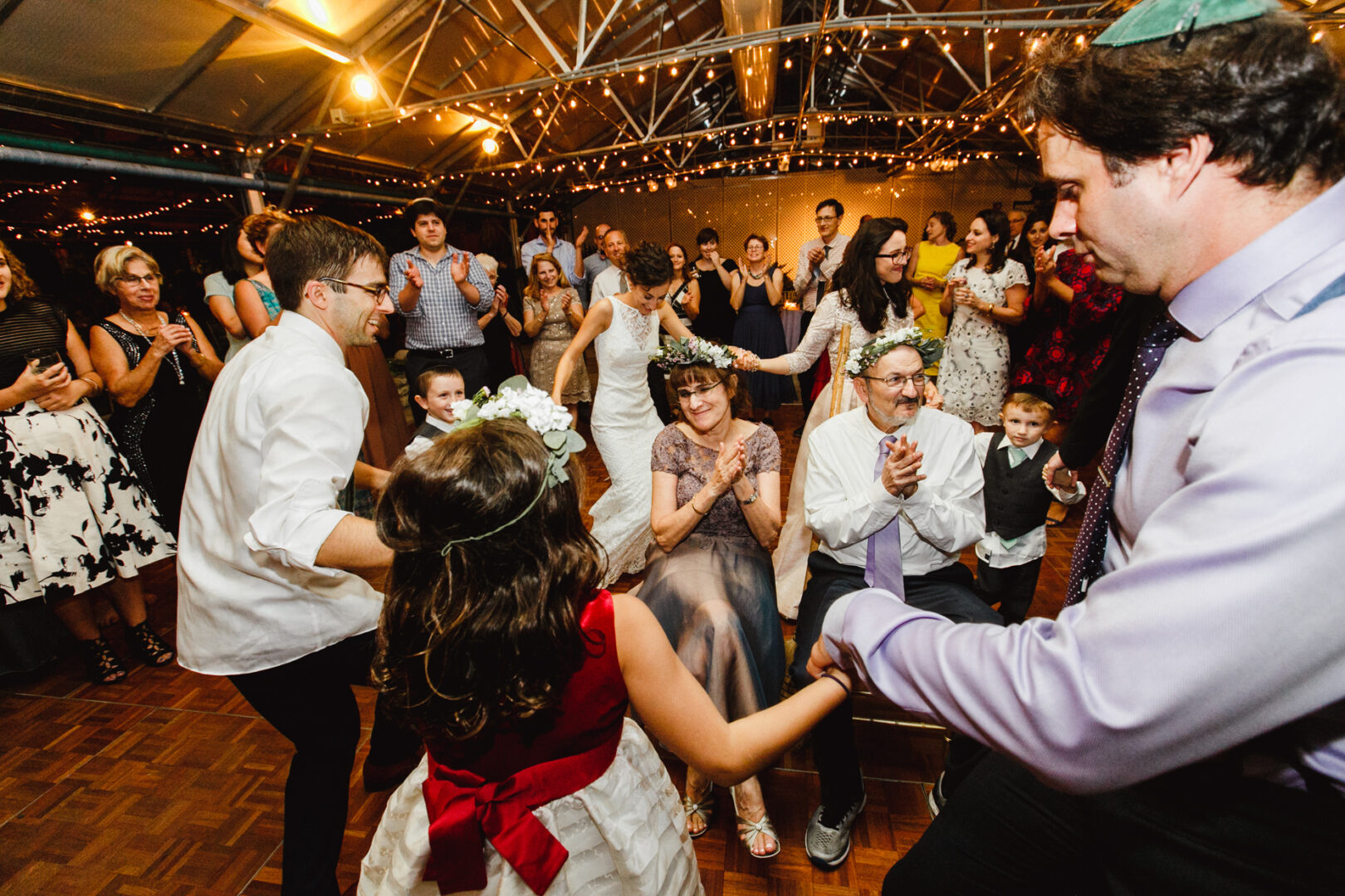 At a Horticultural Center wedding, people dance in a circle at a lively indoor celebration. Some don floral crowns, and the room glows with string lights, creating a magical atmosphere.