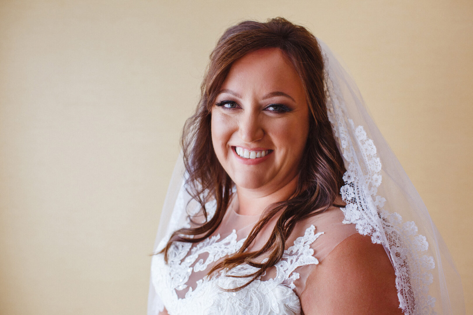 A woman in a white wedding dress and veil smiles at the camera, embodying the charm of a rustic camp-themed wedding against a plain background.