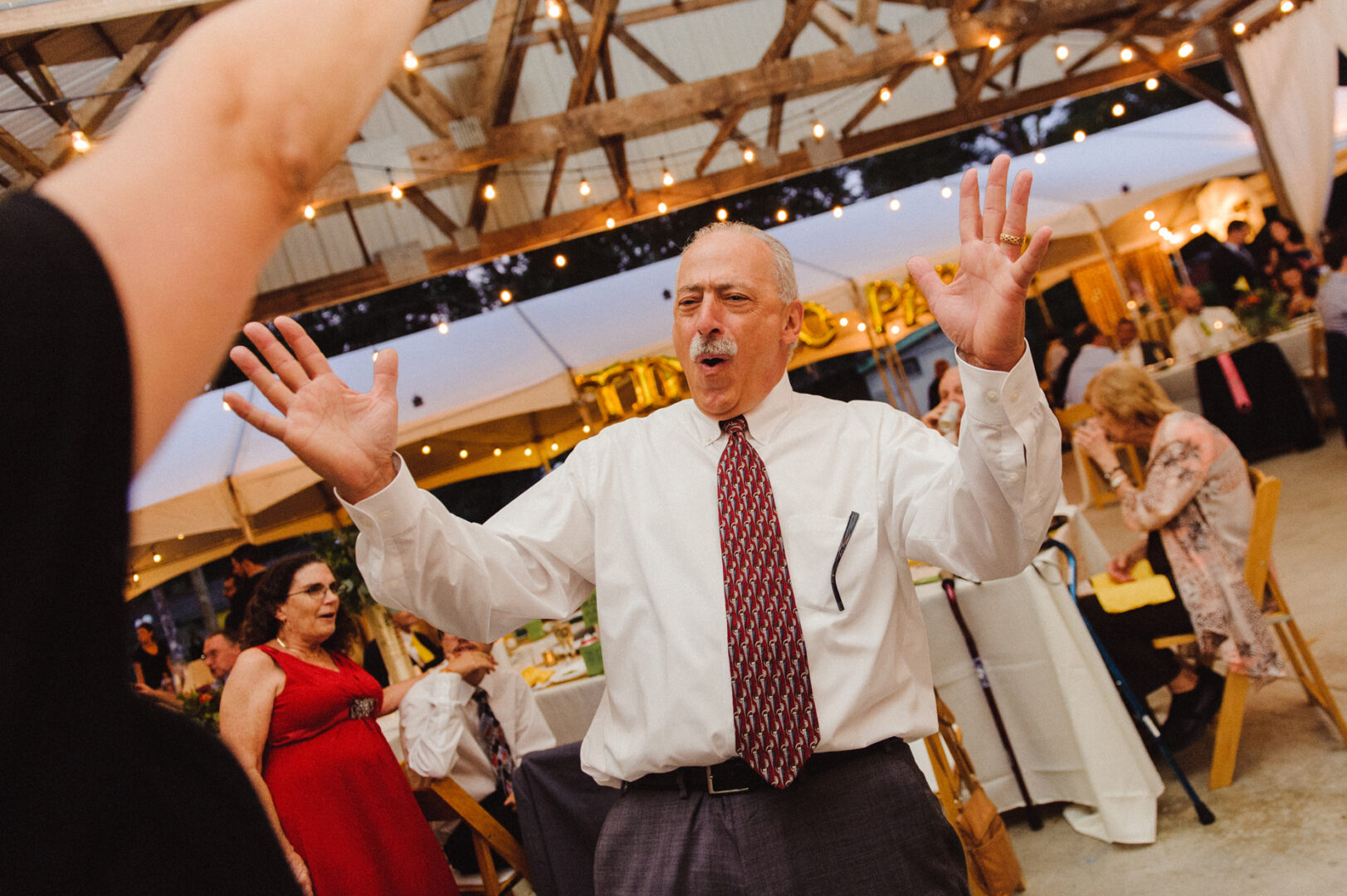 At a rustic camp-themed wedding, a man in a white shirt and tie energetically dances at an indoor event, while people sit at tables in the background.