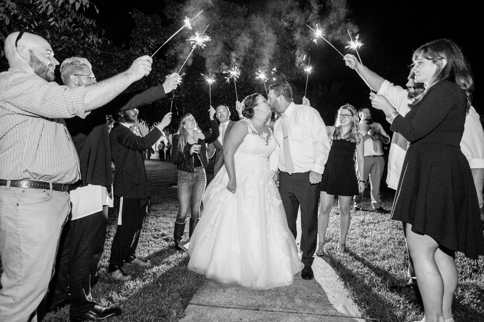 A newlywed couple kisses under a tunnel of sparklers, capturing the magic of their rustic camp-themed wedding. The bride glows in her gown, and the groom is dapper in his shirt and tie. Guests smile and celebrate under the stars, embracing the charming outdoor ambiance.