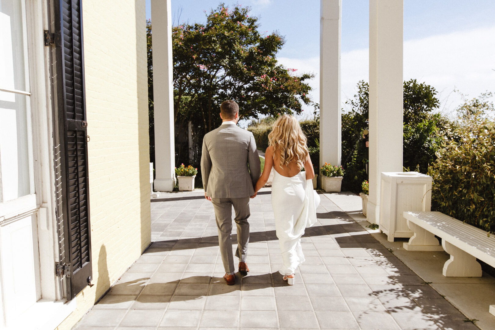 A couple in formal attire walks hand in hand down a sunlit outdoor corridor with columns at Congress Hall, surrounded by lush greenery, capturing the essence of an enchanting Cape May wedding.