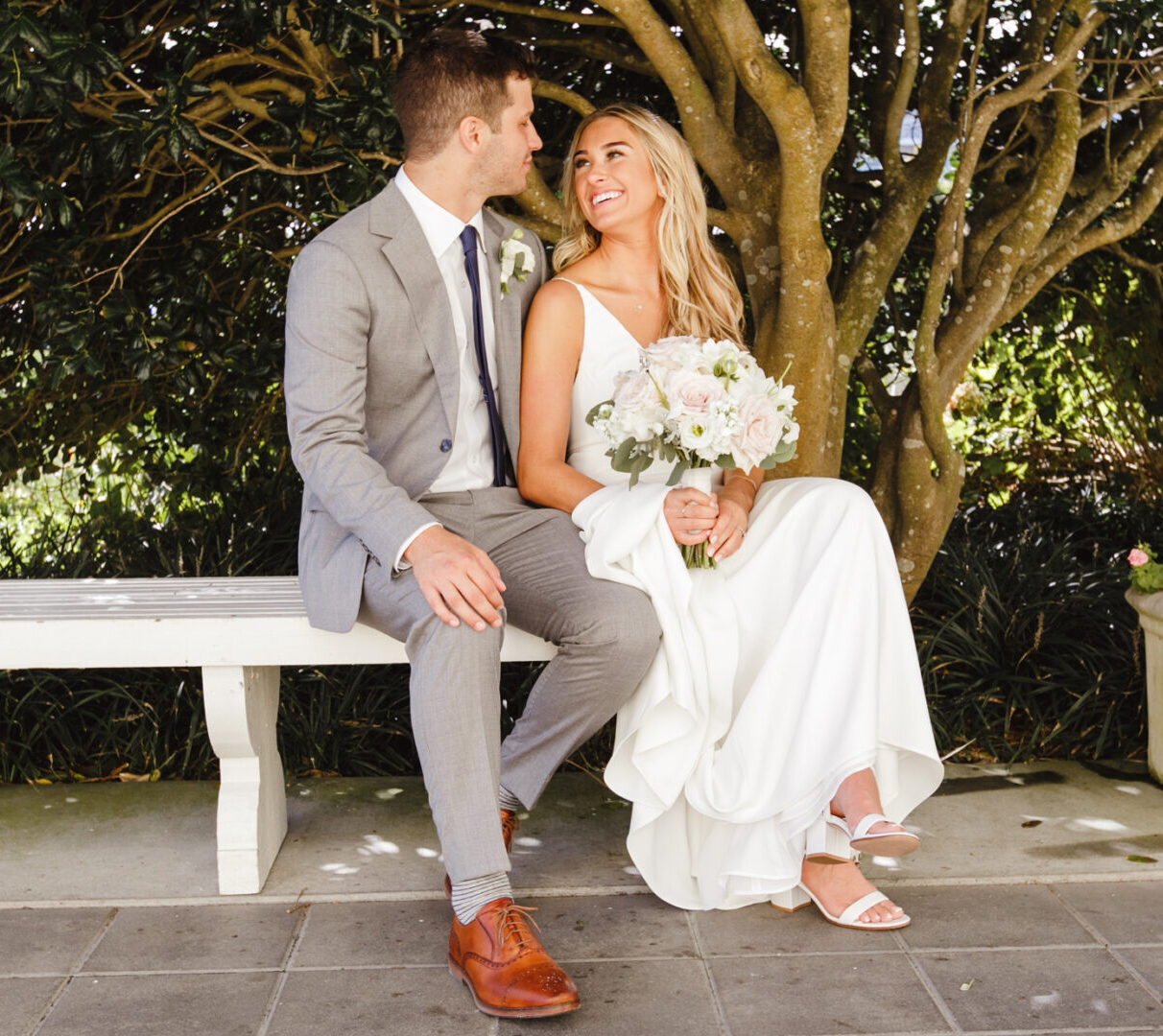 The bride and groom sit on a bench under a tree, exchanging smiles at their Congress Hall Cape May wedding. She holds a bouquet while he sports a gray suit and brown shoes. Nearby, a planter bursts with pink flowers, perfectly complementing their joyful day.