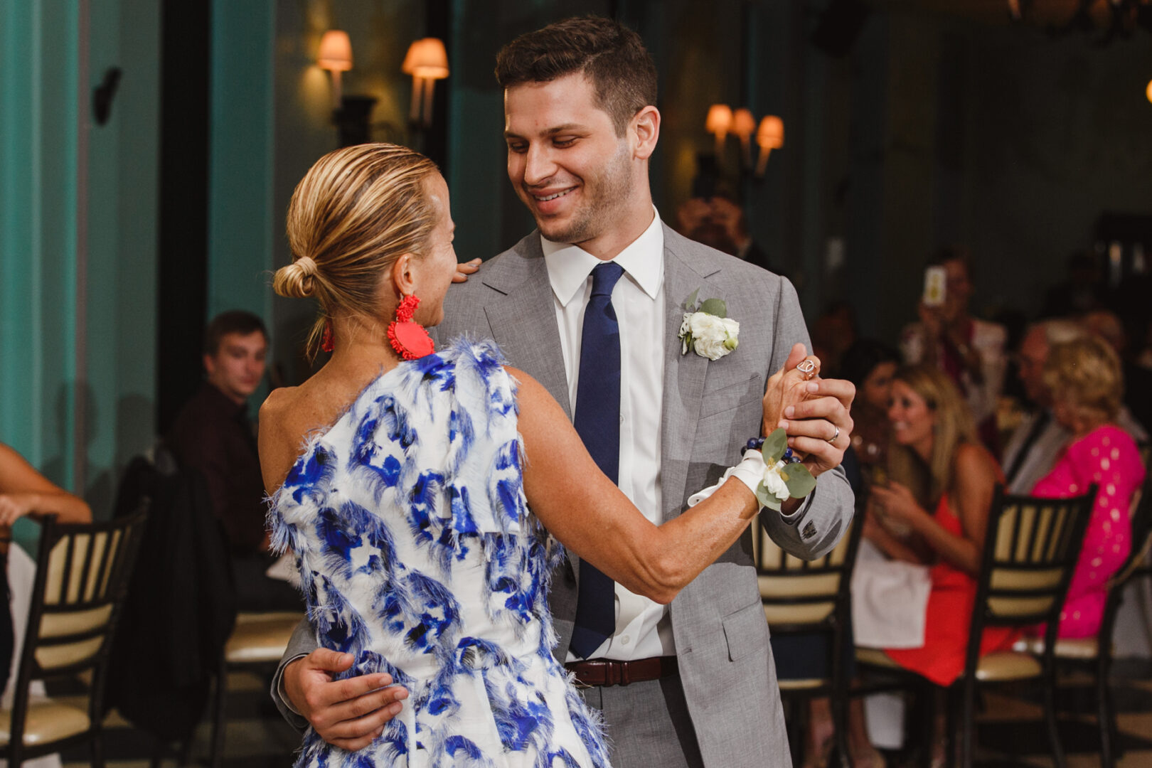 At a charming Congress Hall Cape May wedding, a man in a gray suit gracefully dances with a woman in a blue and white dress. Seated guests and elegant chandeliers twinkle softly in the background, adding to the enchanting indoor atmosphere.