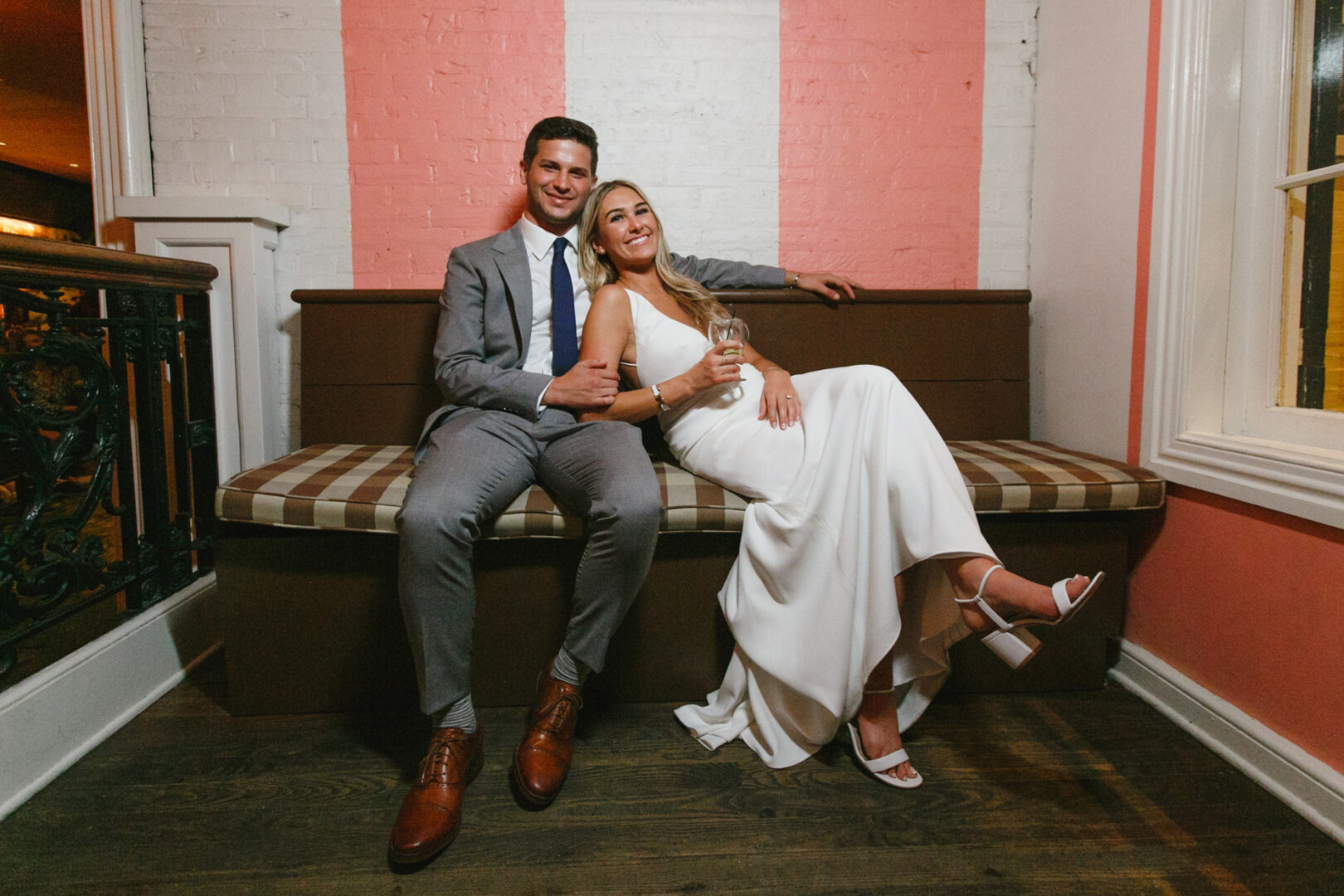 A couple sits on a bench at Congress Hall, Cape May; the man in a gray suit and brown shoes, the woman in a white dress and heels, holding a glass—a perfect moment from their wedding day.