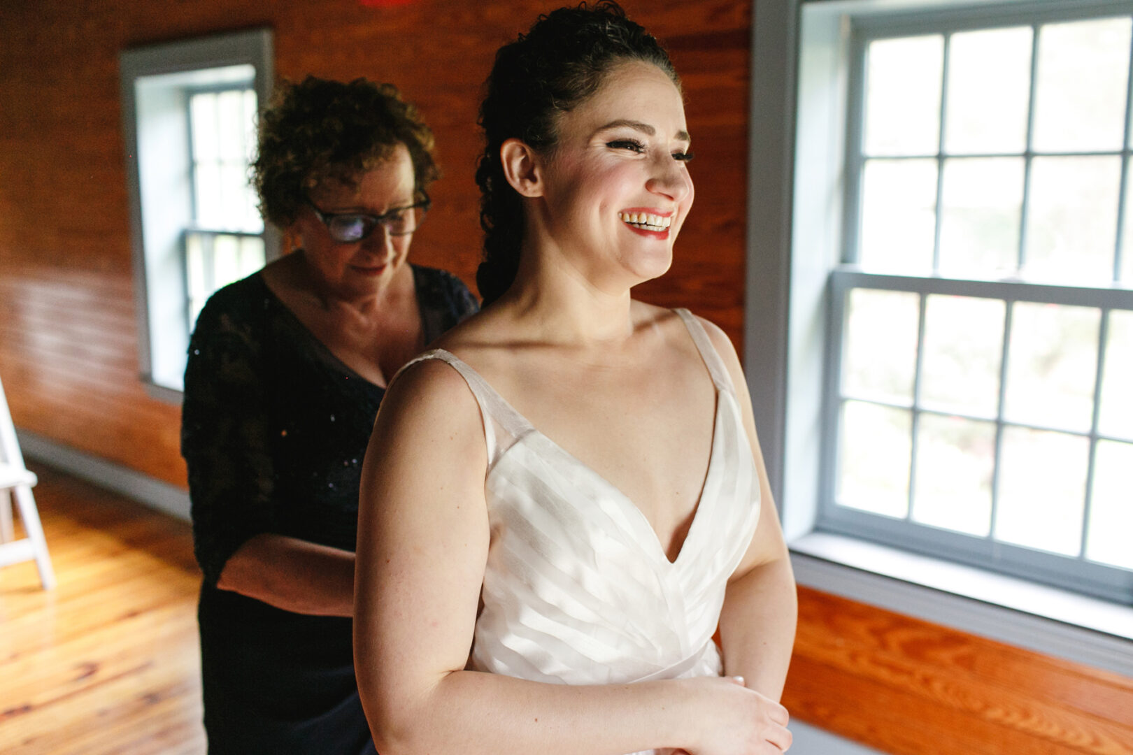 A smiling bride in a white dress stands near a window at the Rockwood Park Carriage House, while another woman assists with the dress in a room with wooden walls.